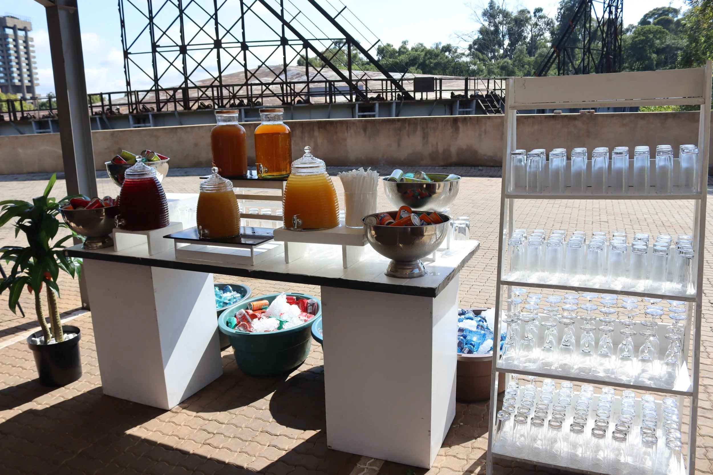 A beverage station setup outdoors with juice dispensers, glasses on a rack, bowls of canned drinks, and a bucket of ice, with a building and trees in the background.