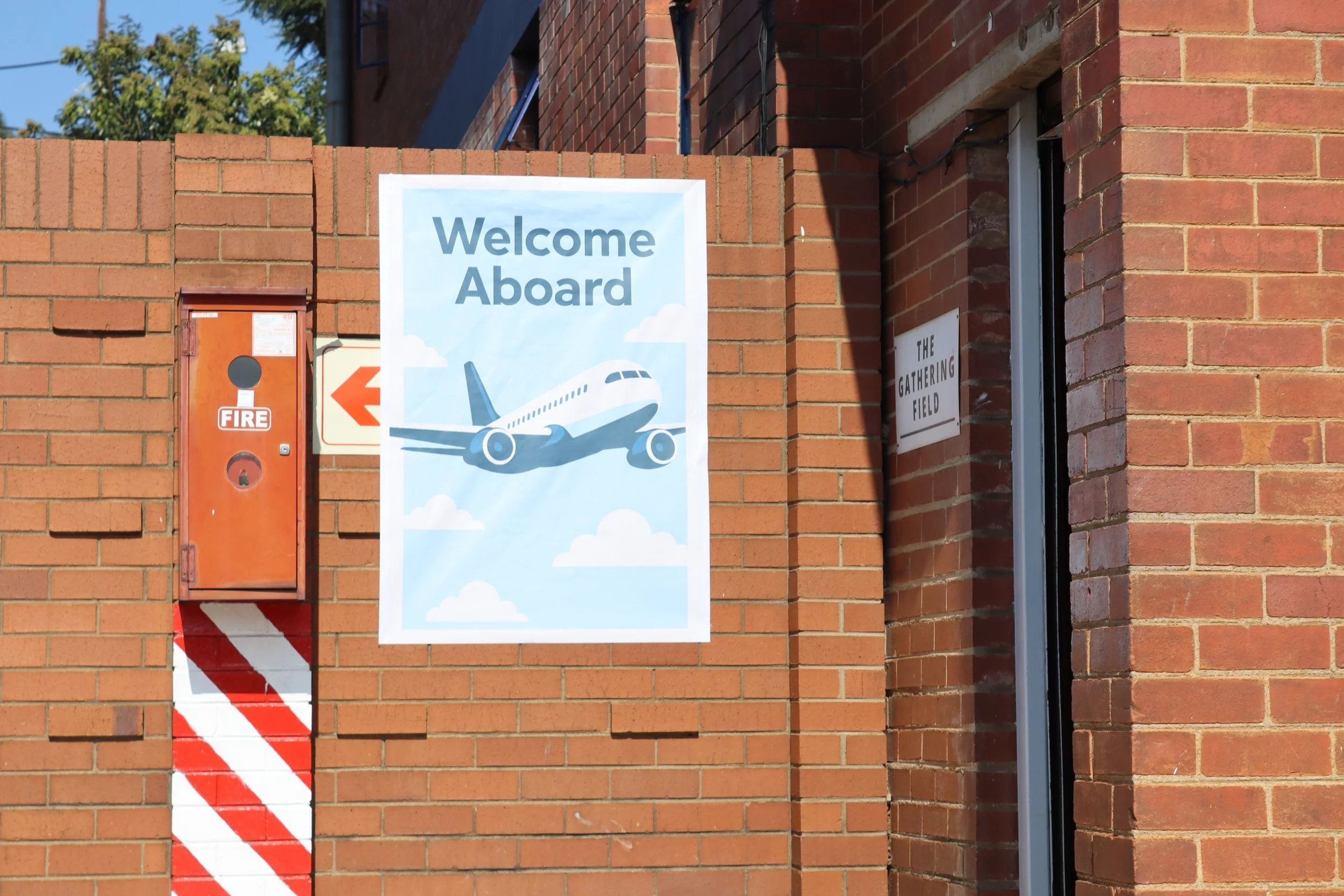 A brick wall with a sign that says "Welcome Aboard" and an illustration of an airplane flying among clouds. There is also a red fire alarm box on the wall.