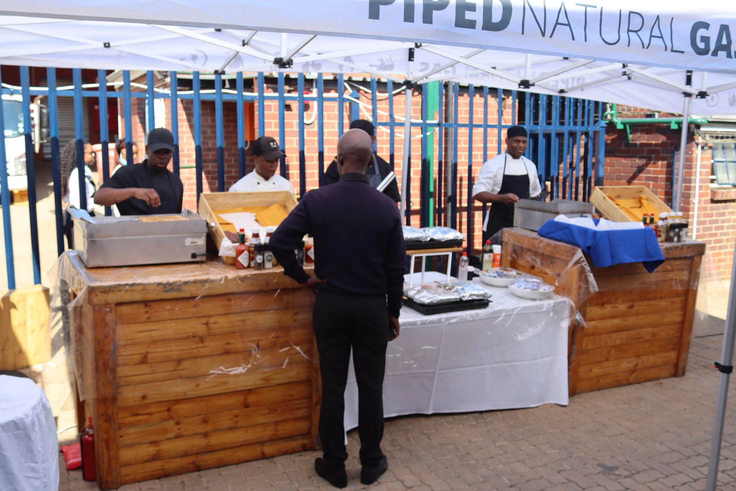 People at a market stall under a white canopy, with grills, condiments, and food items, and a brick building and blue metal fence in the background.
