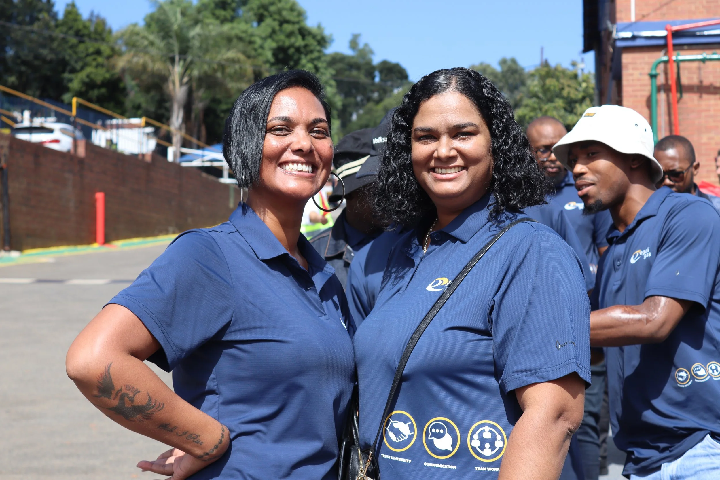 Two women smiling and standing outdoors in a line of people wearing matching blue shirts, with trees and a brick building in the background.