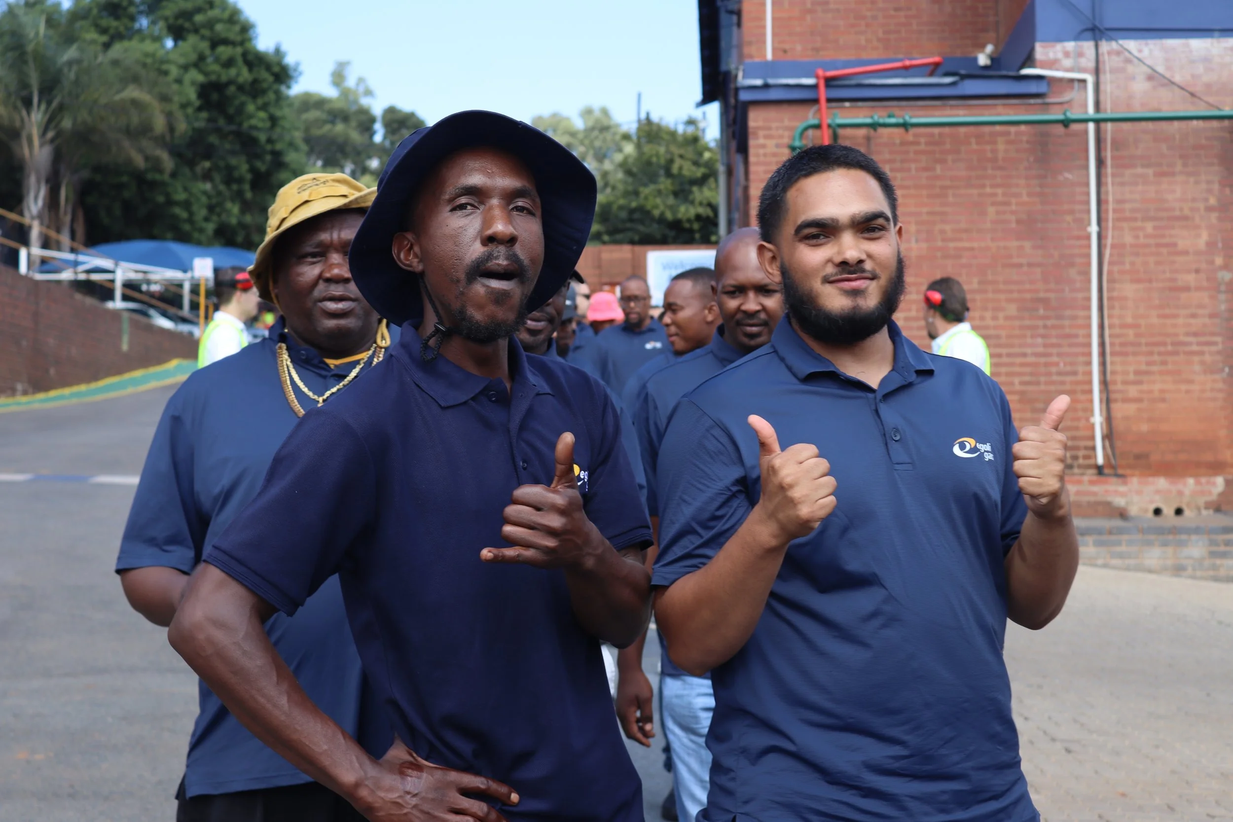 Group of men outdoors, wearing blue shirts, some with thumbs up, standing near a brick building and trees.