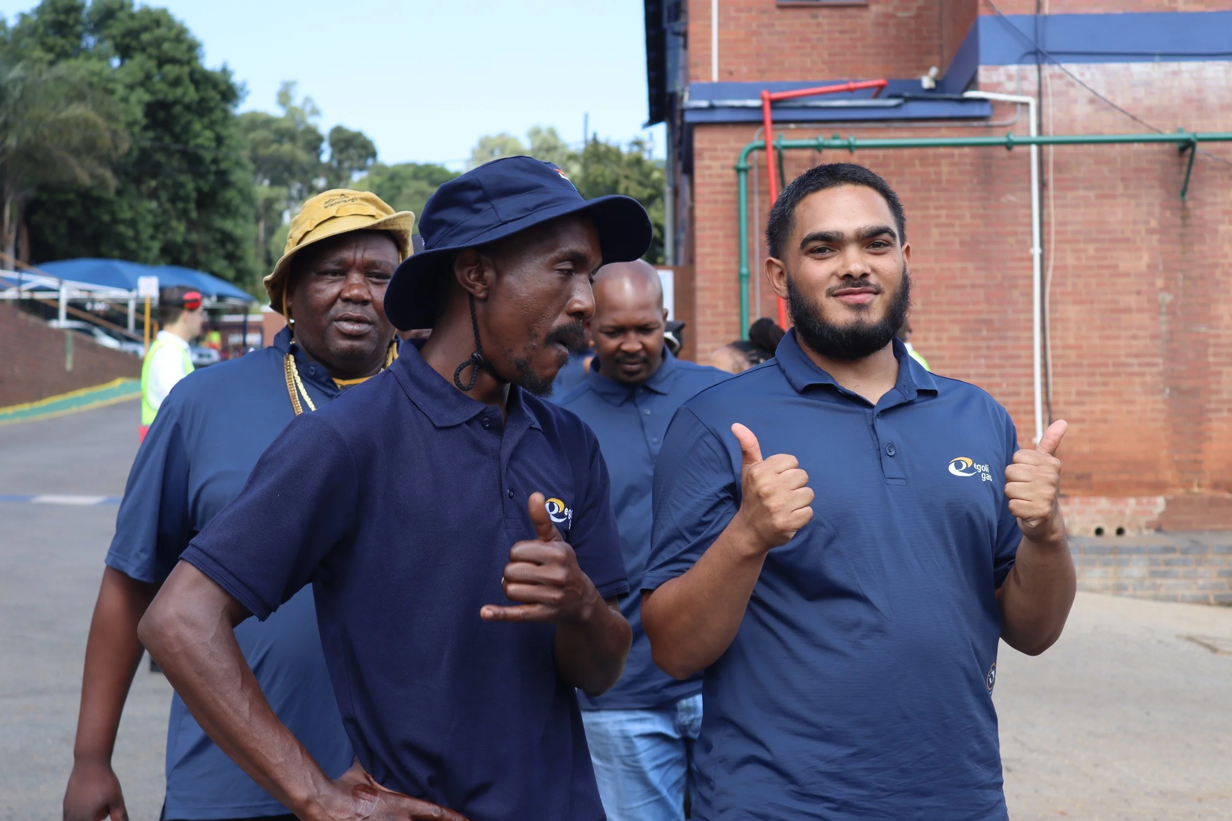 Three men outdoors, two of them making thumbs-up gestures, wearing blue shirts with a logo, in front of a brick building and green trees.