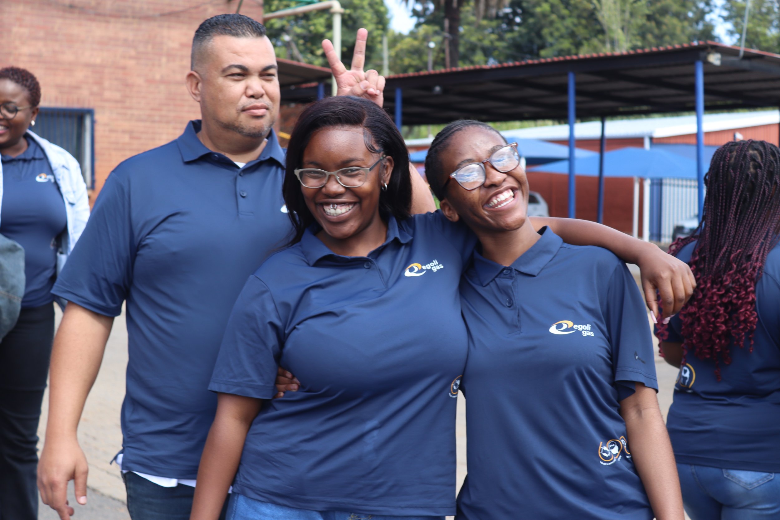 Group of people wearing blue shirts with a logo, smiling, and having fun outdoors, with one person making a peace sign behind another person.