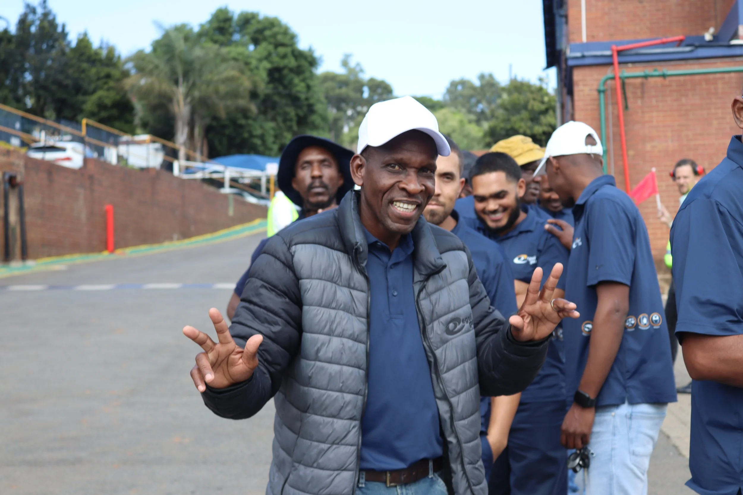 A group of men outdoors, with one man in the foreground smiling and making a peace sign with his fingers, wearing a white cap and a gray vest.