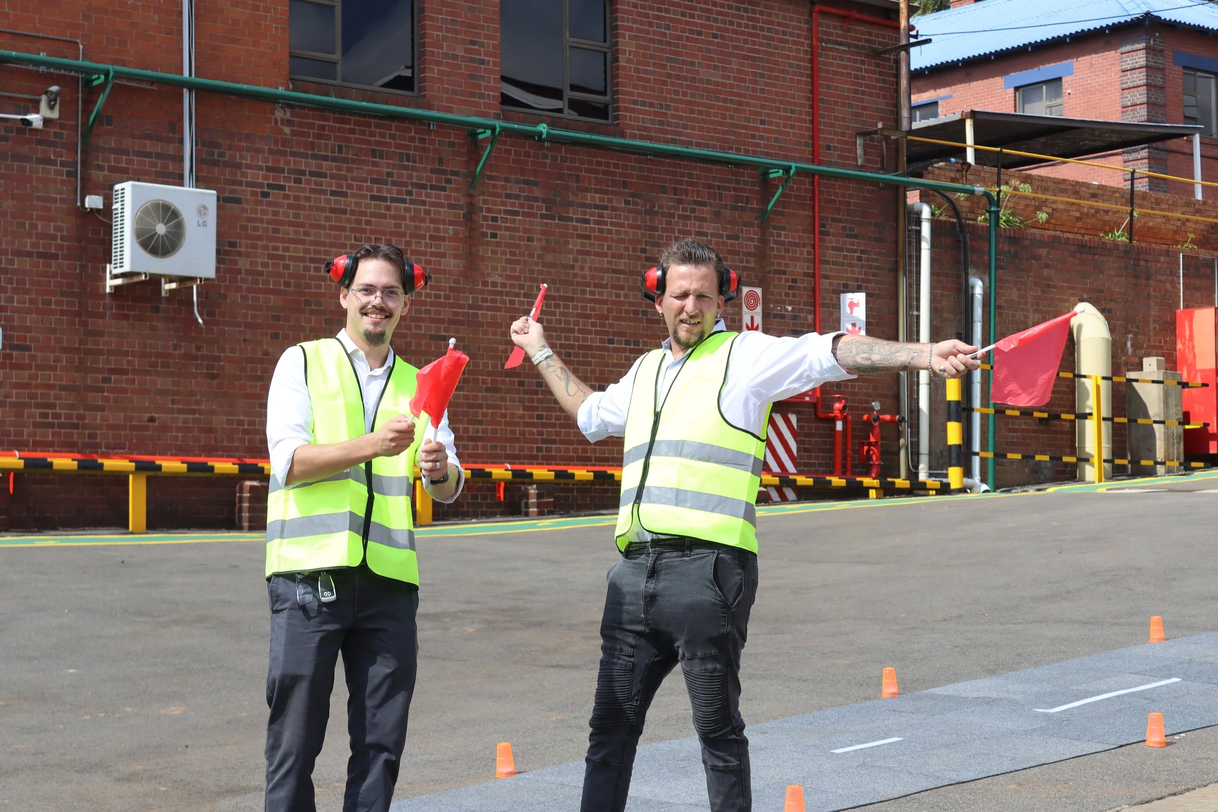 Two men wearing high visibility vests and hearing protection standing outdoors near a brick building, holding red flags and orange cones are set up on the ground with more cones arranged in a line.
