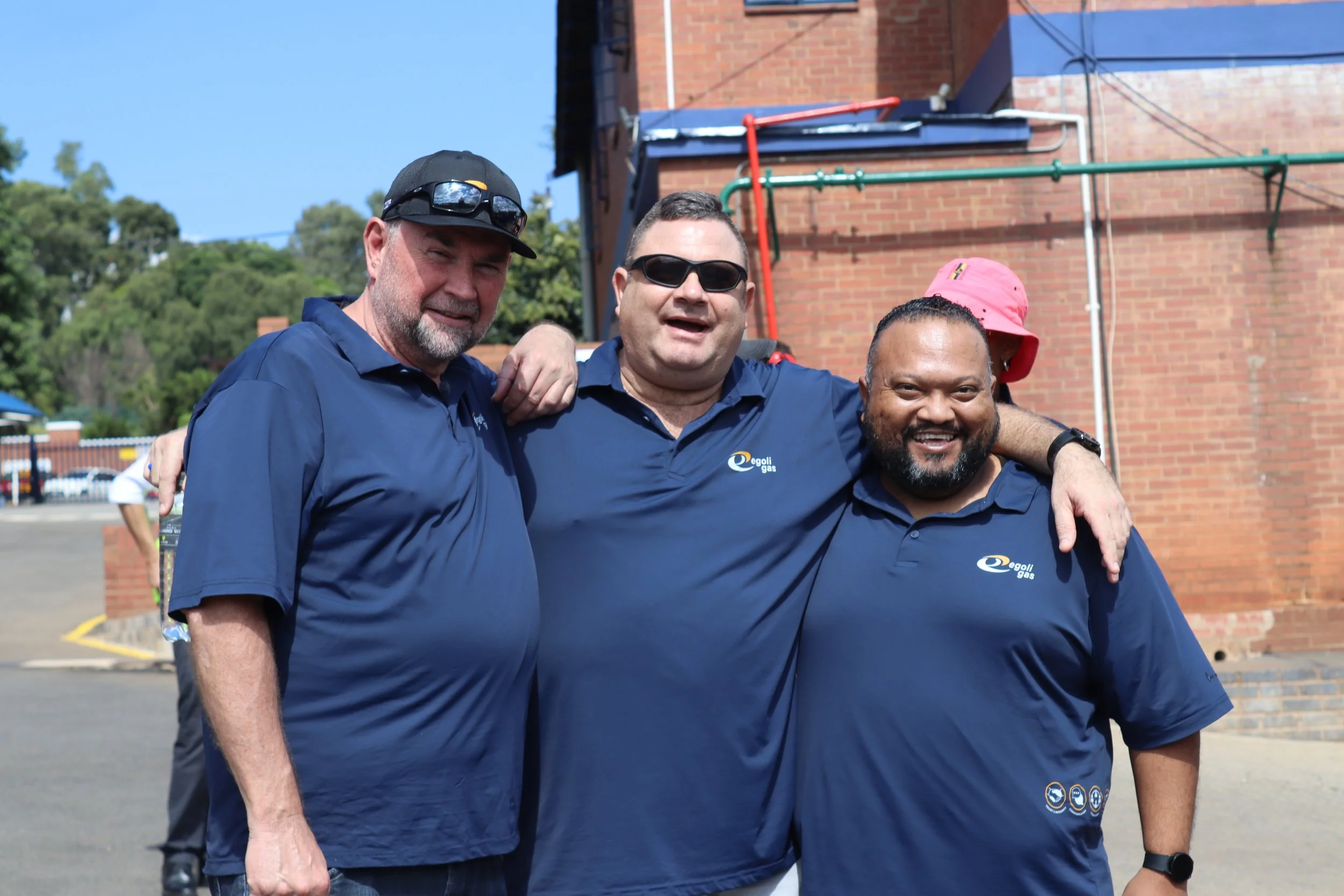Three men smiling and standing close together outdoors, wearing blue shirts with a logo, with a brick building and trees in the background.