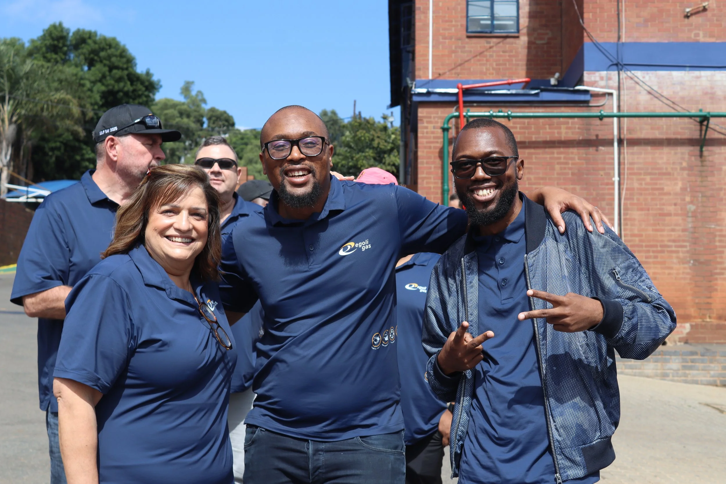 A group of people smiling and posing outdoors, some wearing matching blue shirts with a logo, in front of a brick building and green trees on a sunny day.