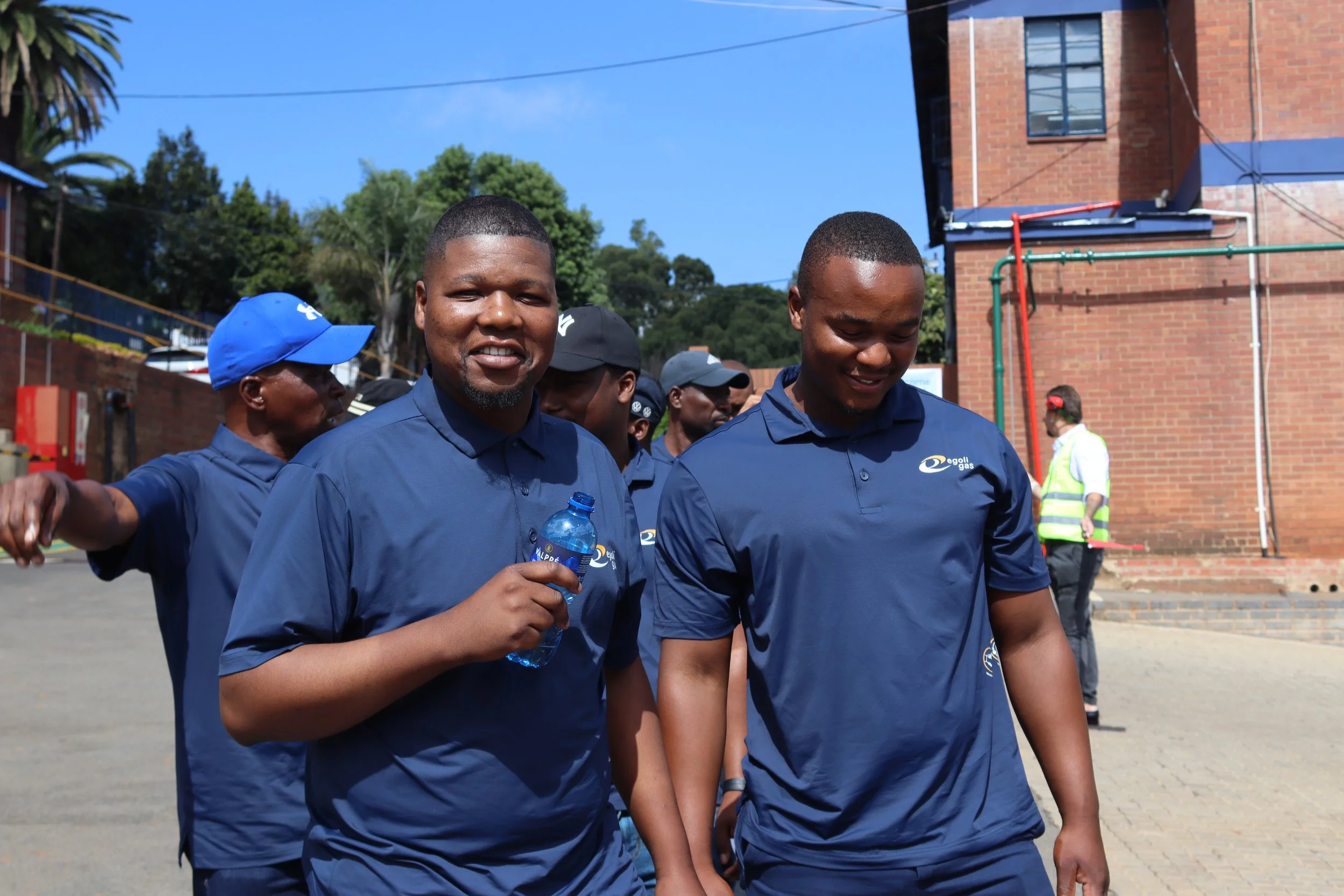 Group of men in blue shirts walking outdoors on sunny day, some holding water bottles, with brick building and trees in background.