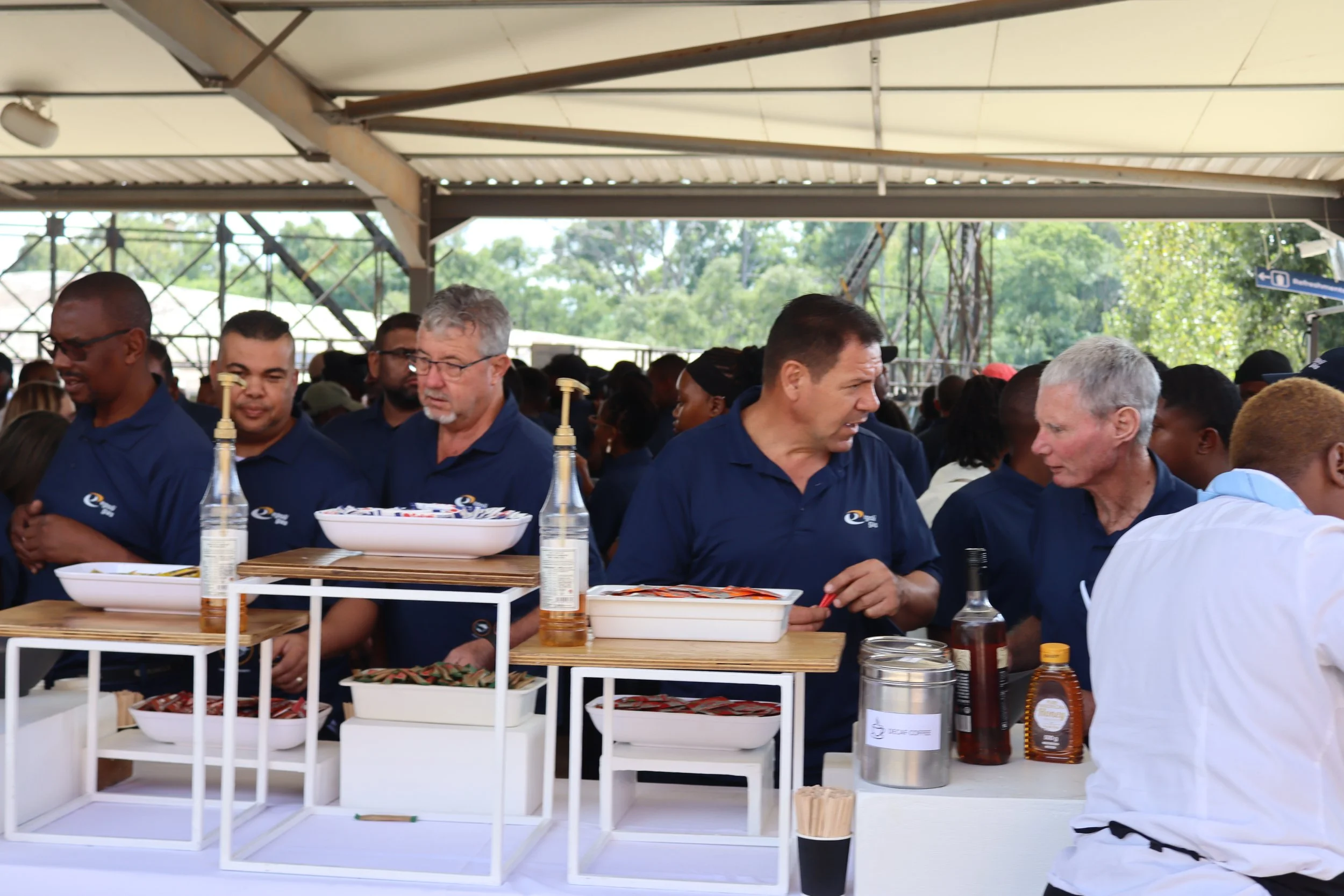 Group of people wearing dark blue shirts serving food at an outdoor event under a canopy, with tables holding bowls, bottles, and condiments.