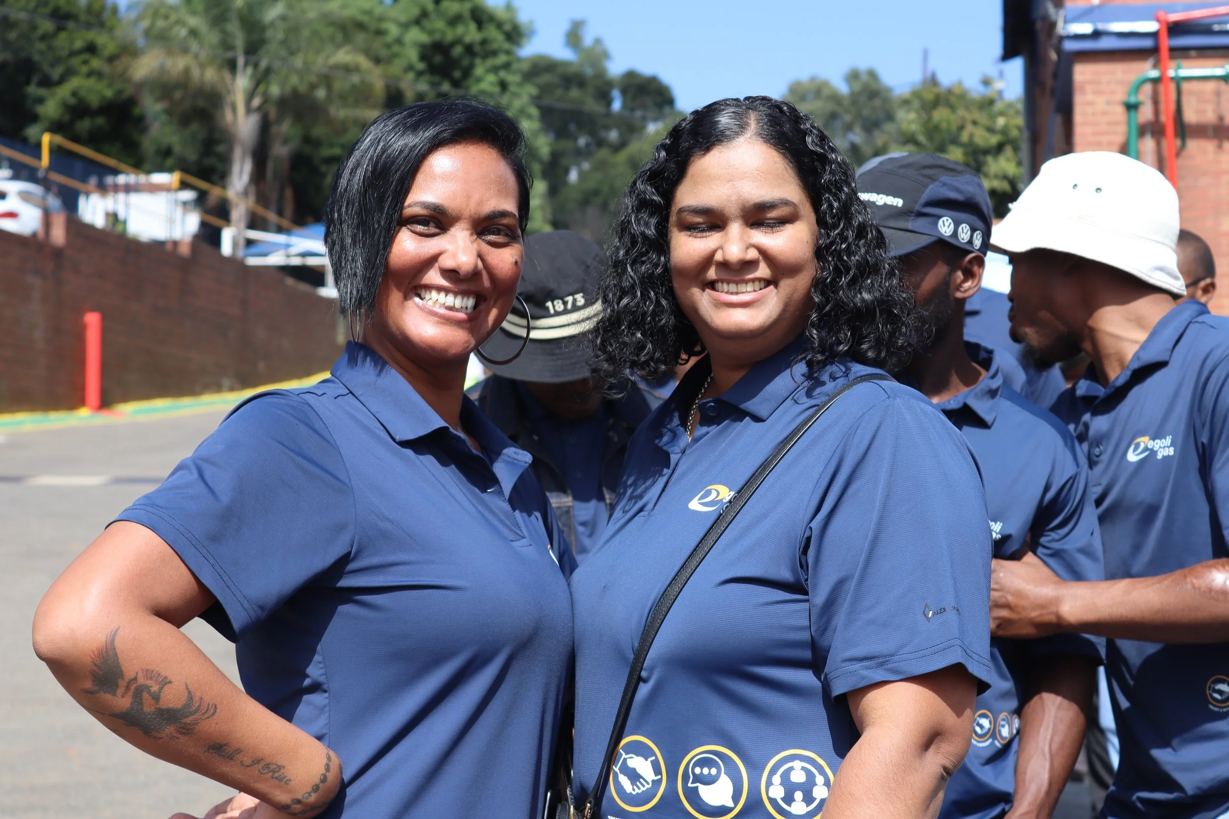 Two women wearing blue shirts smiling outdoors with others in the background.