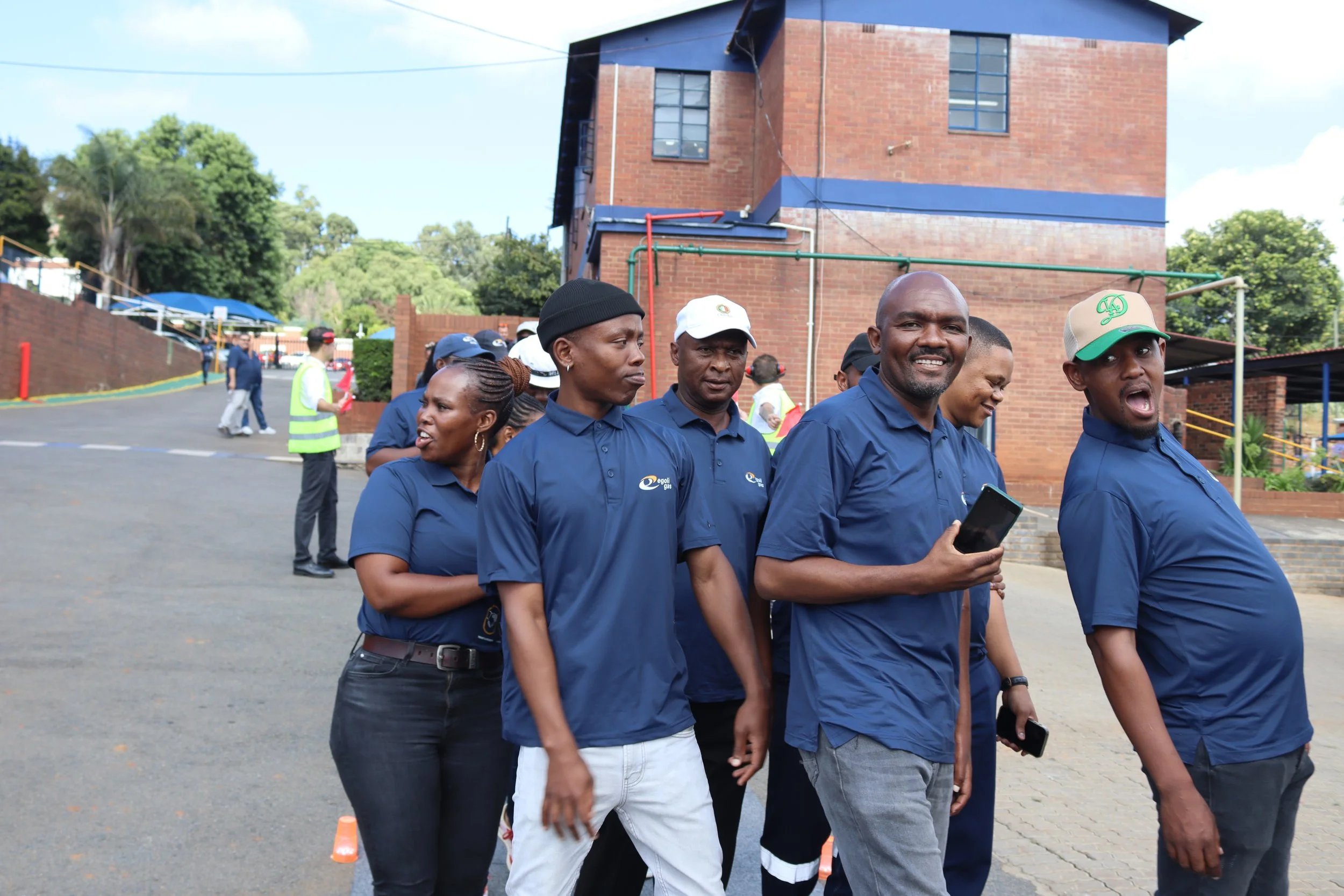 Group of people walking outdoors near a brick building with trees in the background. They are wearing blue shirts, some are smiling, and one person is holding a smartphone.