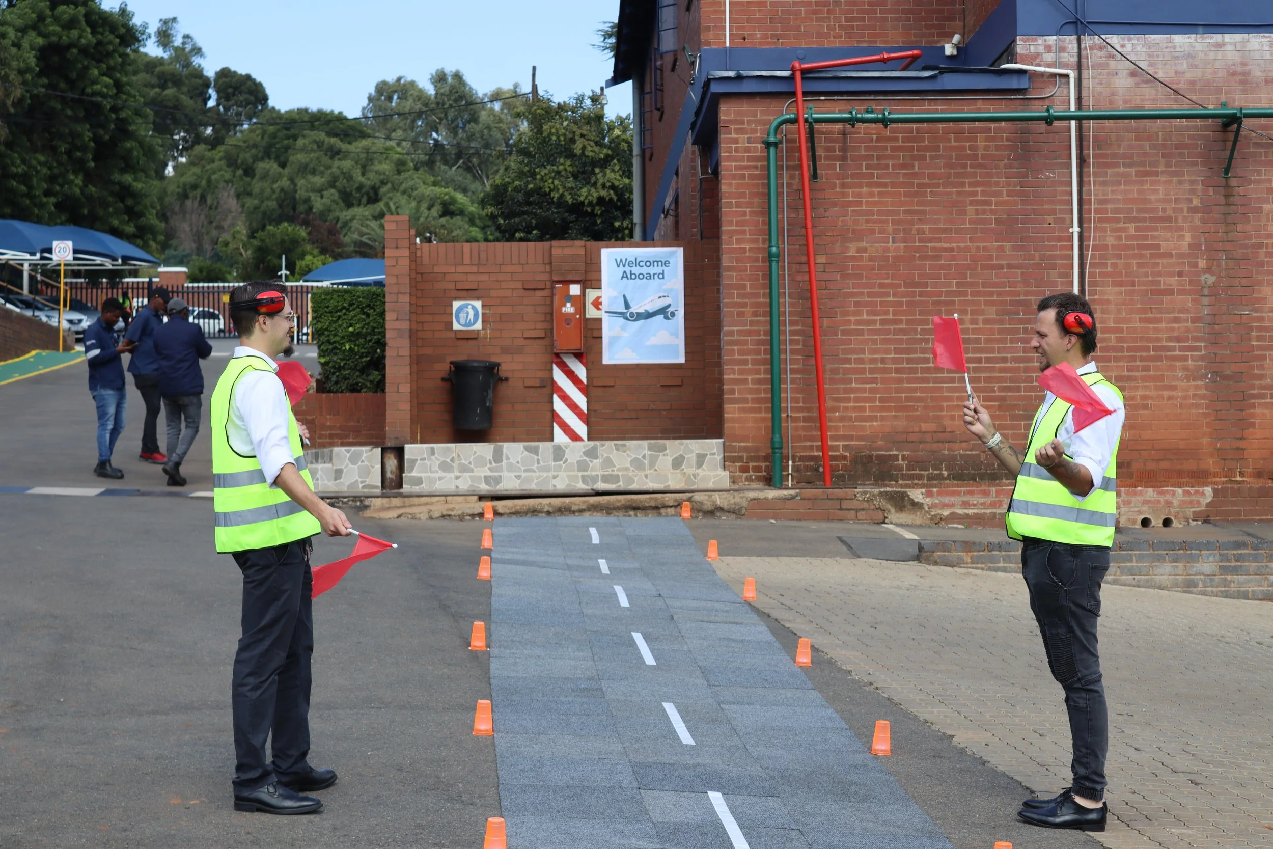 Two men in safety vests and headphones holding red flags are guiding pedestrians and traffic at an outdoor stop sign area near a brick building. Several people are walking in the background, and there are parking lot and trees visible.