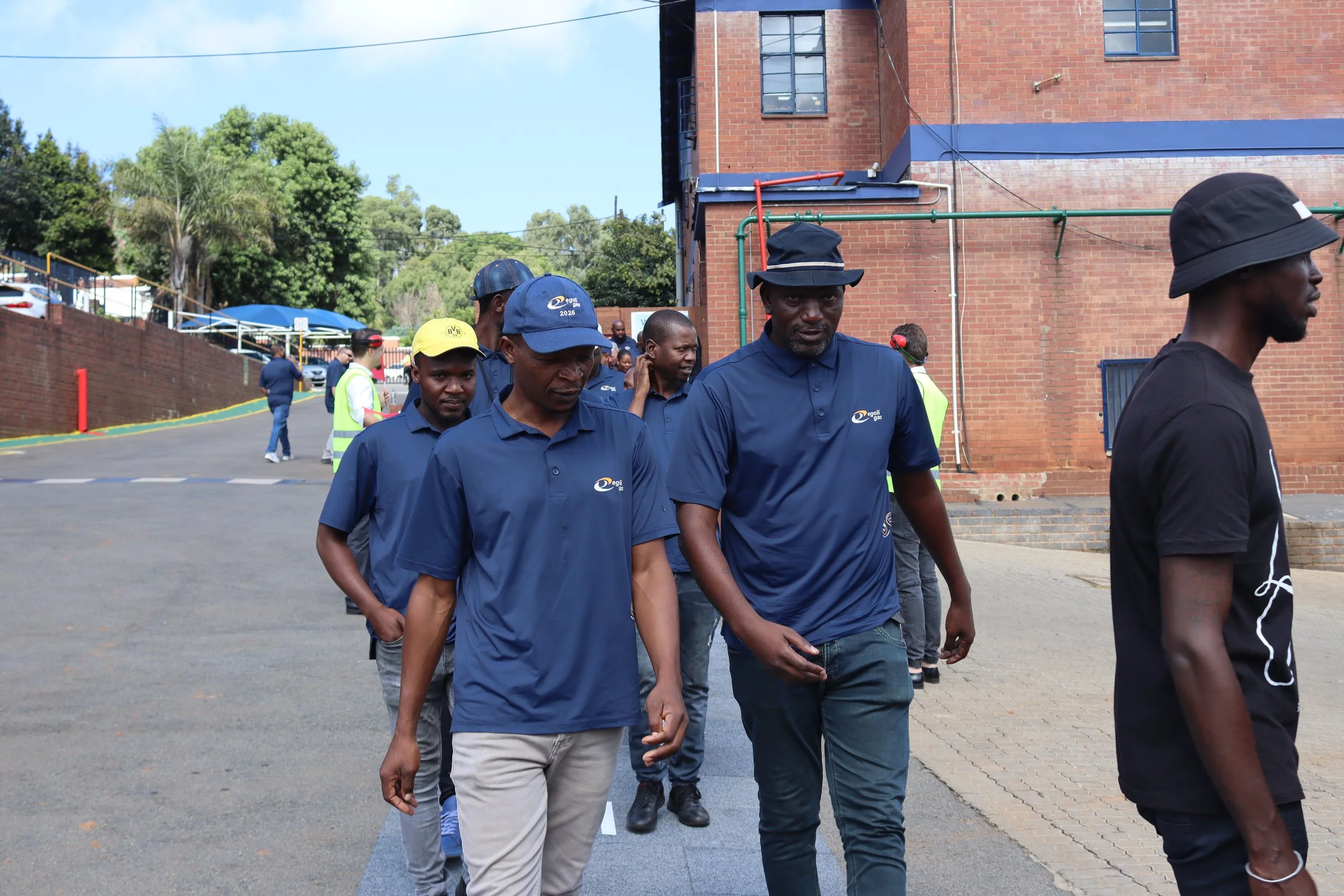 A group of men walking outdoors near a brick building and trees. The men are dressed casually, some wearing hats and shirts with a logo, and the scene appears to be in a commercial or industrial area.