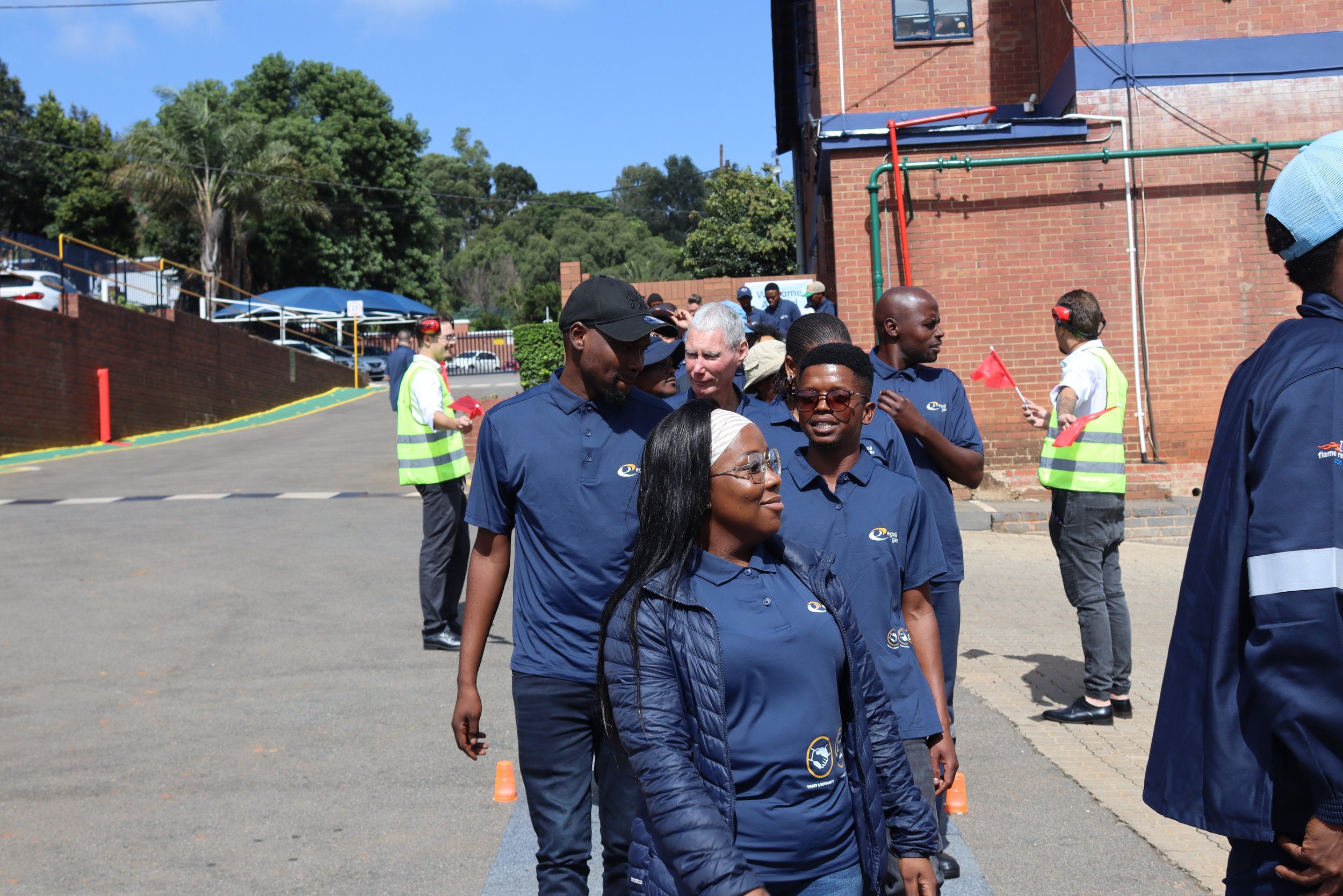 A group of people standing outdoors, wearing blue shirts with a handshake logo, participating in a business or community event. Some are holding red flags, with a brick building, trees, and parked cars in the background.