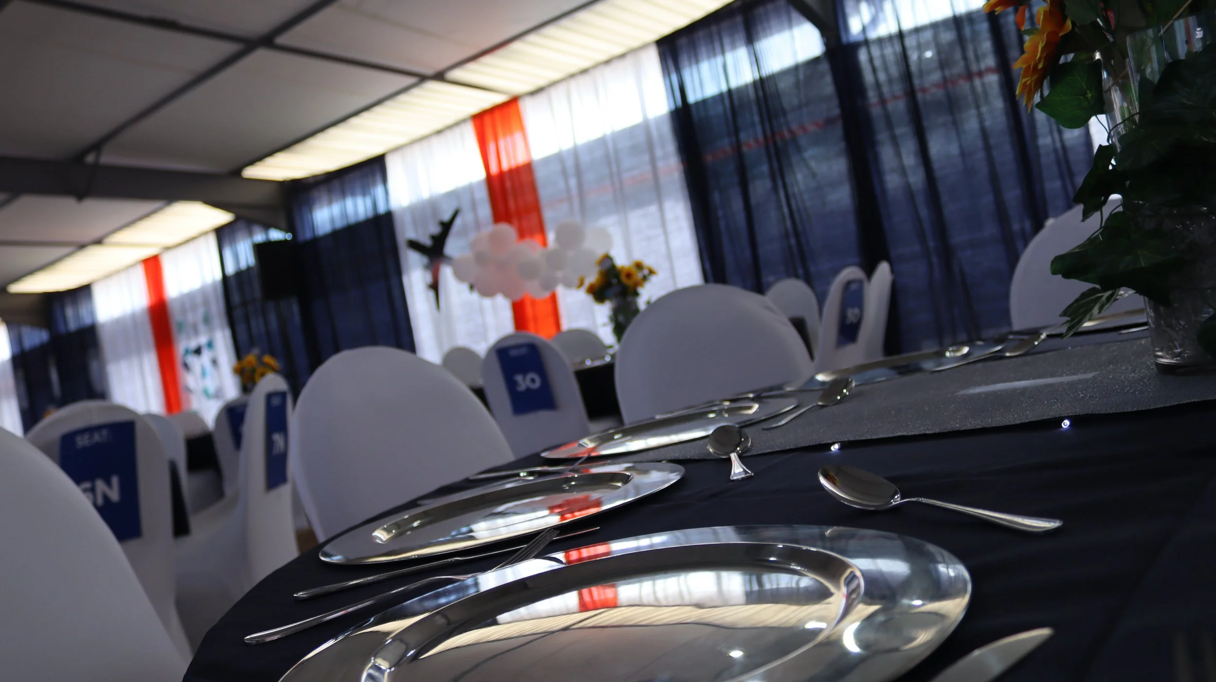 Set dining table with silverware and white chairs at an event with decorated backdrop and balloons.