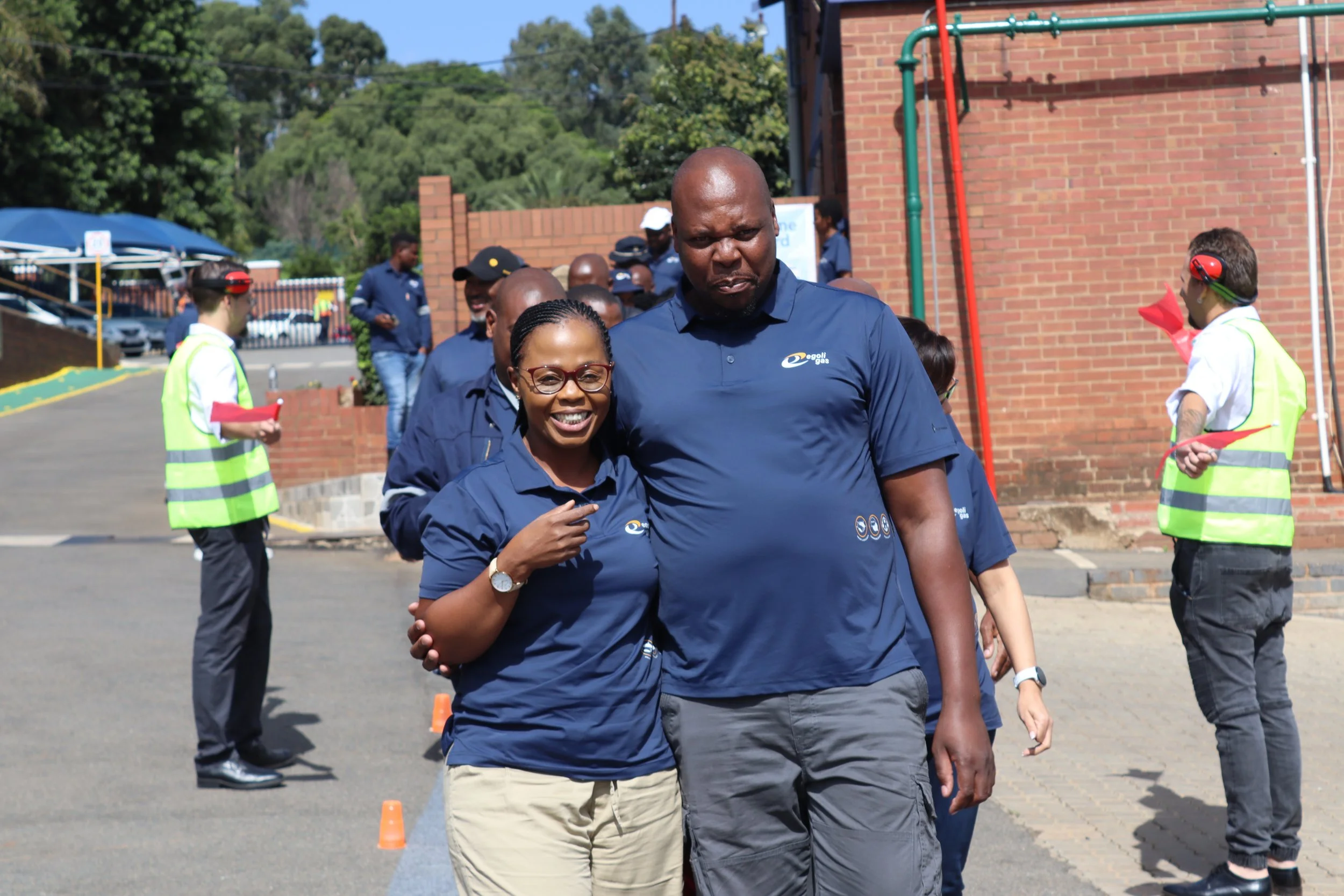 A group of people wearing blue shirts walking outdoors near a brick building with green pipes, some wearing headphones and safety vests, on a sunny day.
