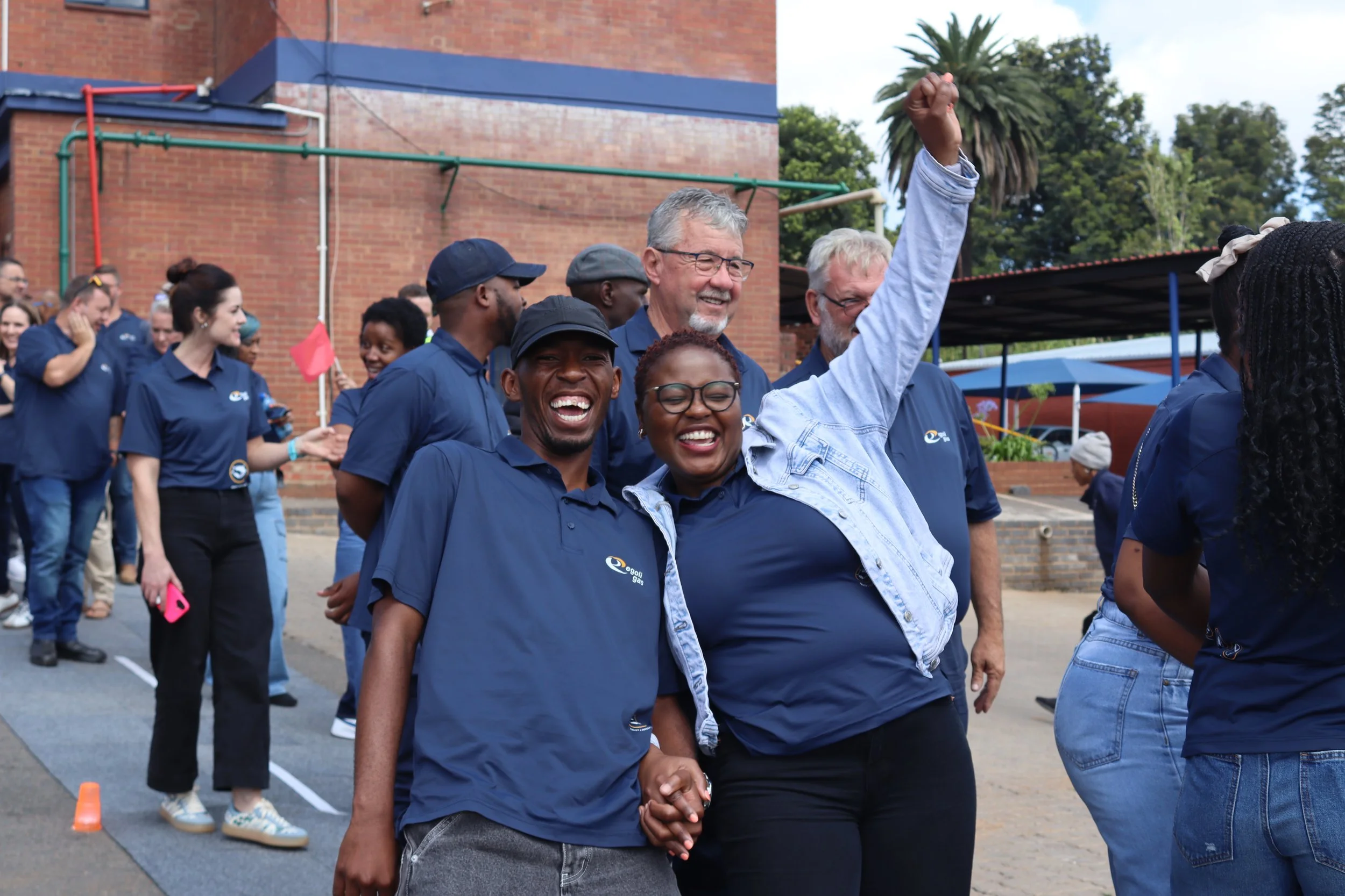 Group of diverse people, smiling and holding hands, walking outdoors during daytime, with some raising a fist in celebration.