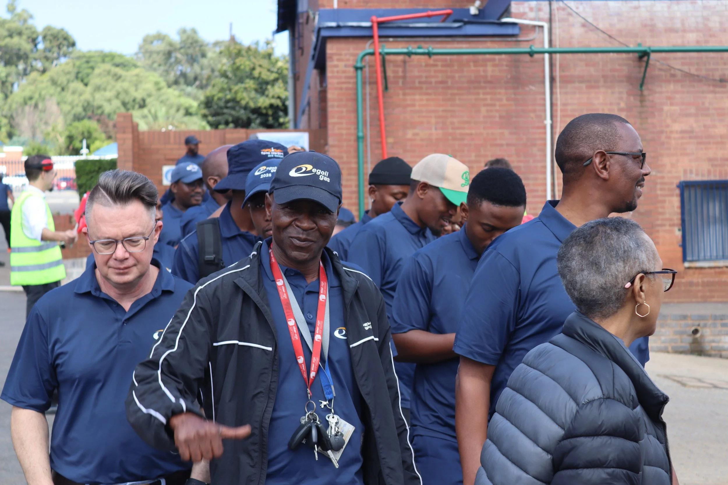 A group of people, mostly men, gathered outdoors in front of a brick building. They are dressed in navy blue shirts, some wearing caps with the logo "egoli gas". Some individuals are smiling or looking downward, and a few are wearing lanyards and key