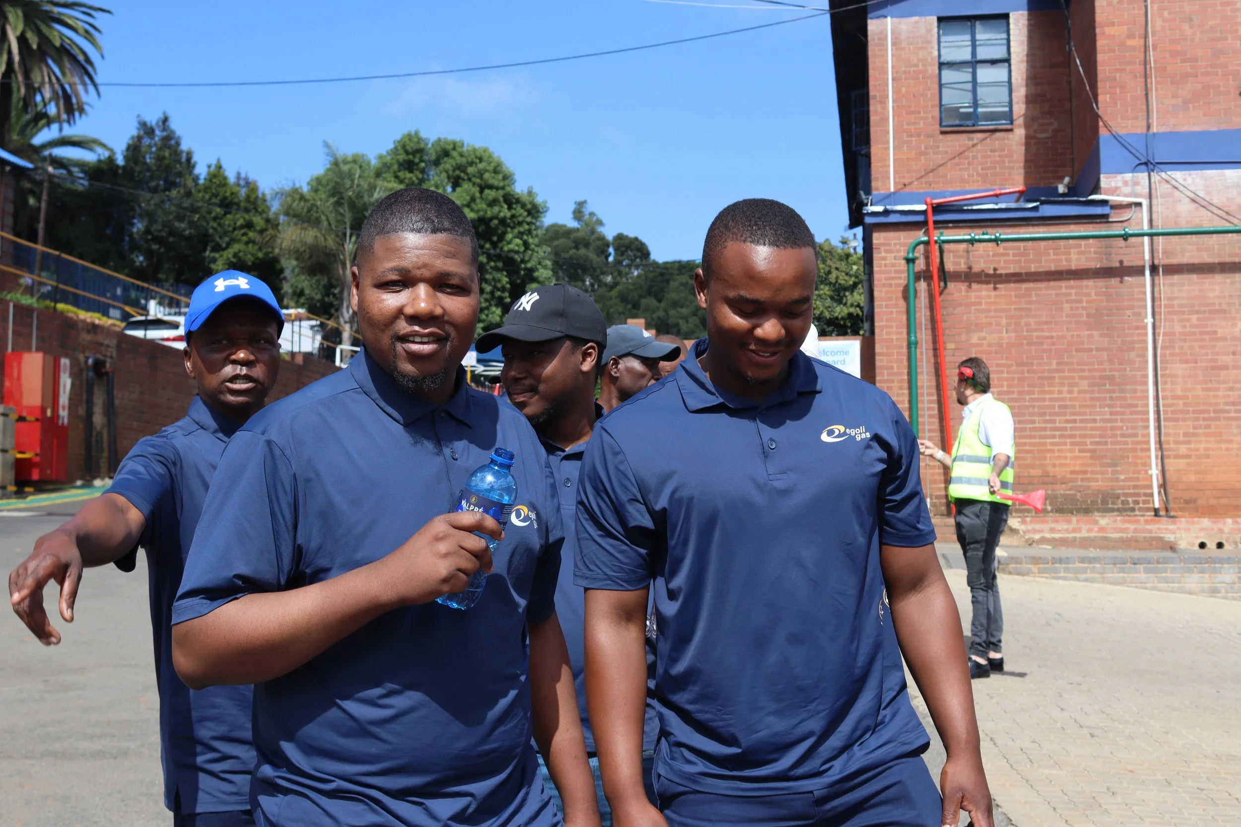 A group of men walking outdoors, wearing matching dark blue shirts, with trees and a brick building in the background. One man is holding a bottle of water.