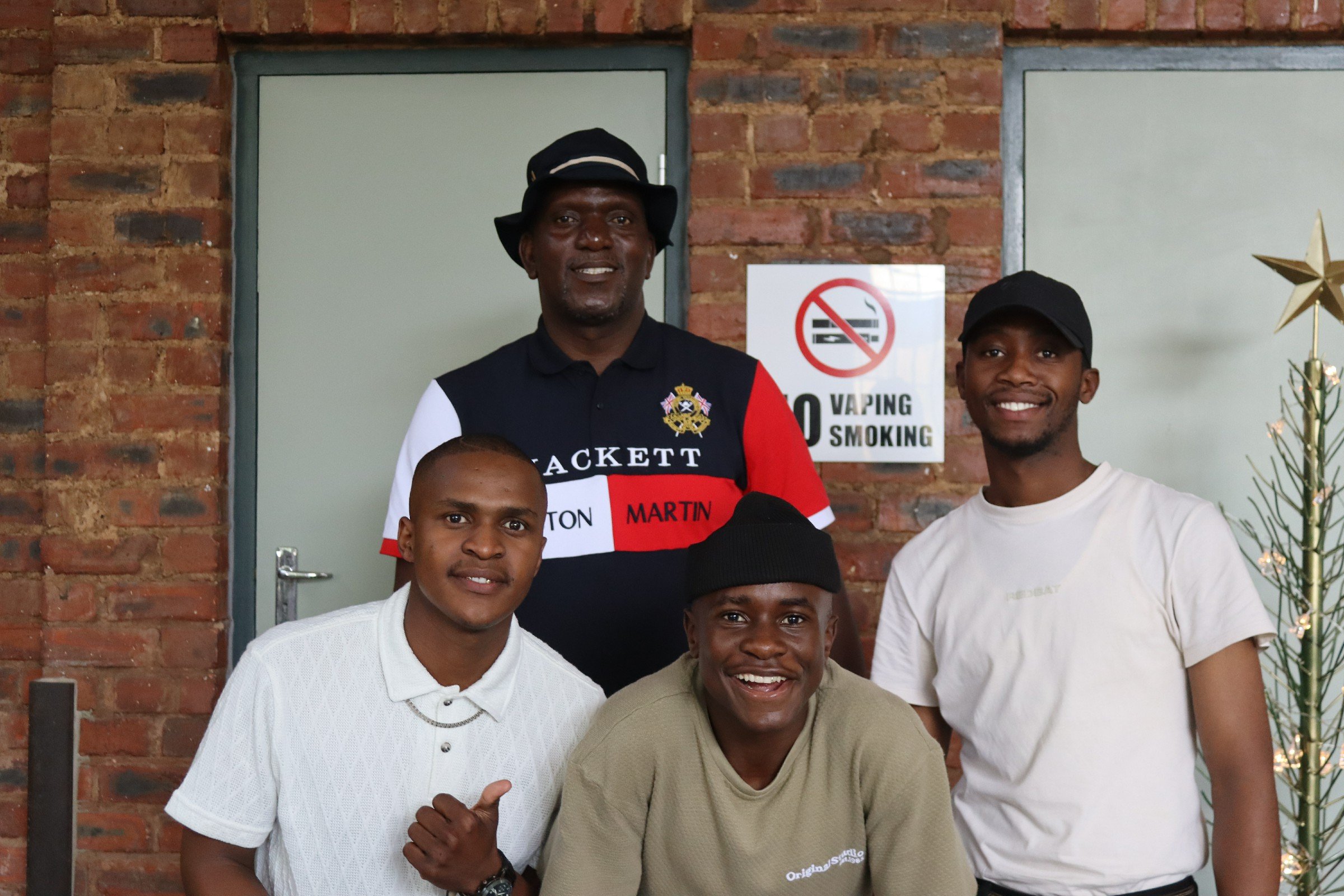Group of five smiling young men standing in front of a brick wall, with a 'No Vaping Smoking' sign and a decorated Christmas tree with stars and lights to the right.