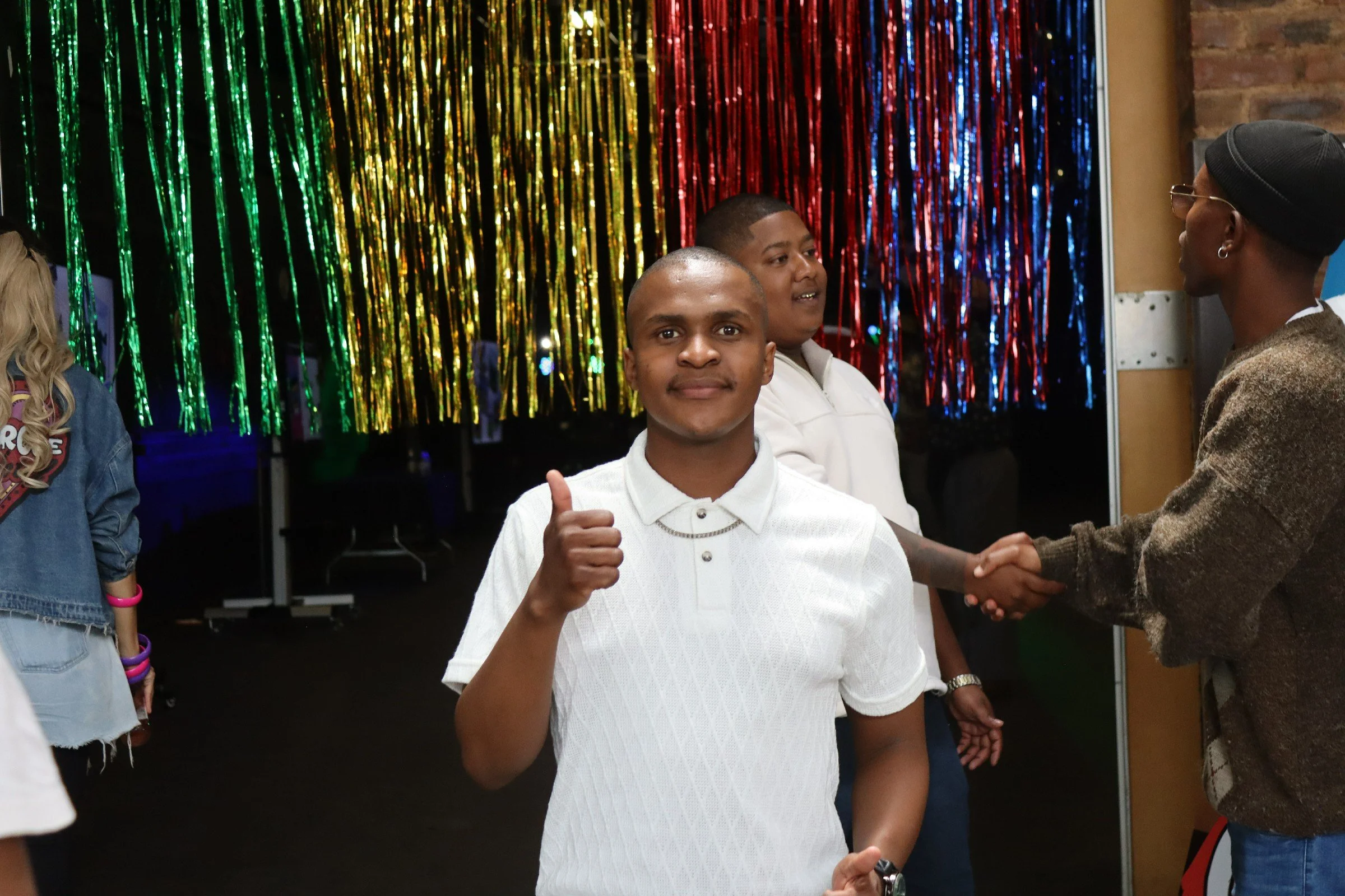 Young man giving a thumbs-up gesture at a party with colorful metallic streamers in the background.