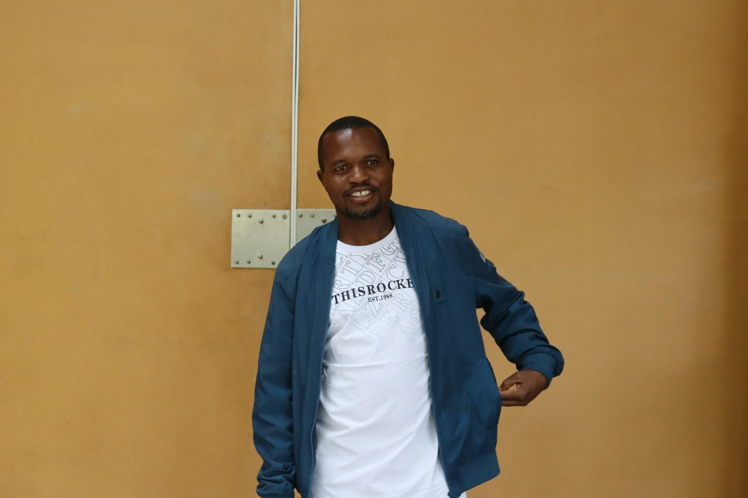 A man with short hair and a goatee, wearing a white graphic T-shirt and a blue jacket, standing against a plain beige wall.