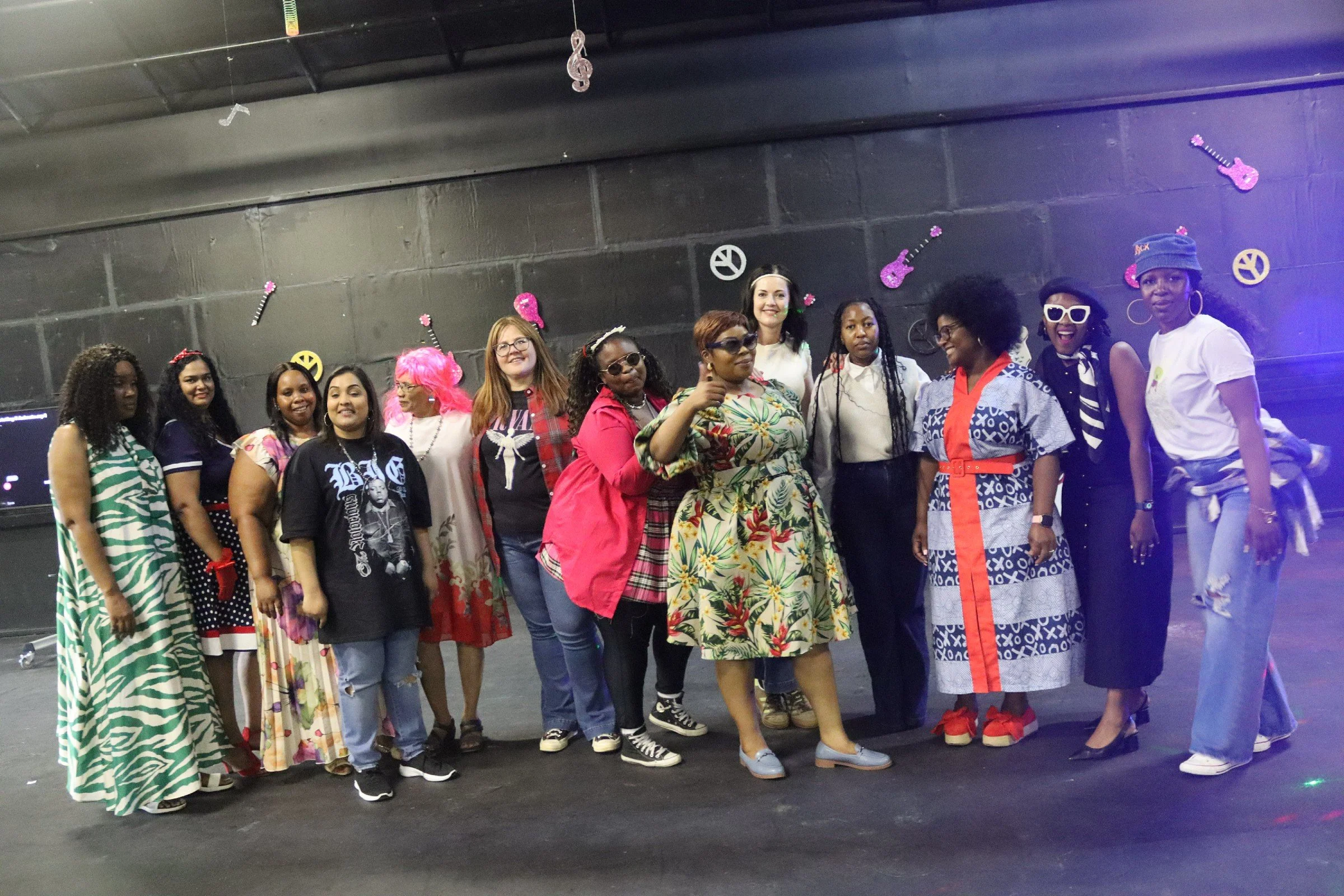 A diverse group of women standing in a line and smiling at the camera against a black wall decorated with pink and yellow guitars and peace signs. They are wearing colorful, patterned, and casual clothing.