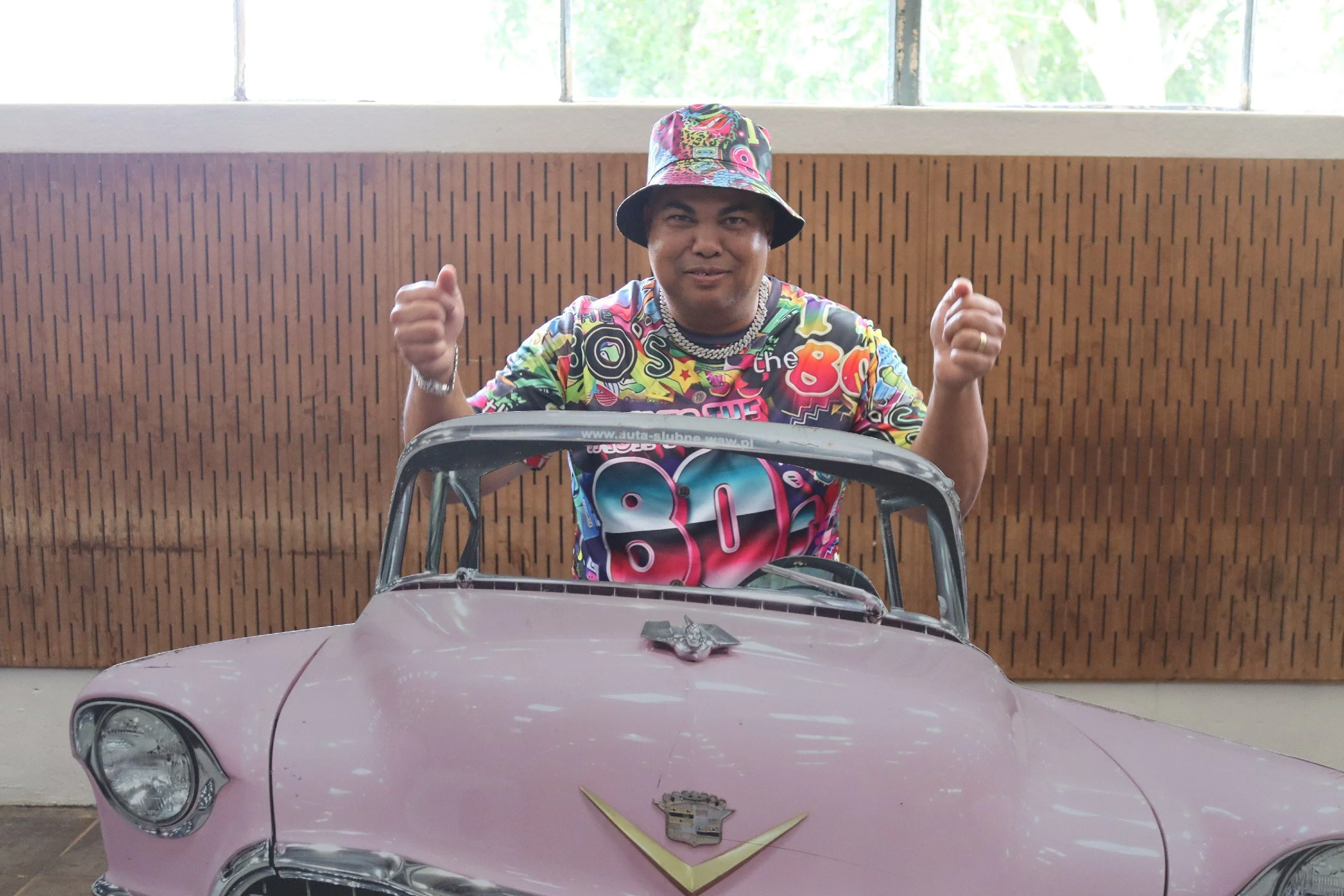 A person wearing a colorful shirt and hat is sitting behind a pink toy car, smiling and making fists with both hands in front of a wooden-paneled wall and large windows.