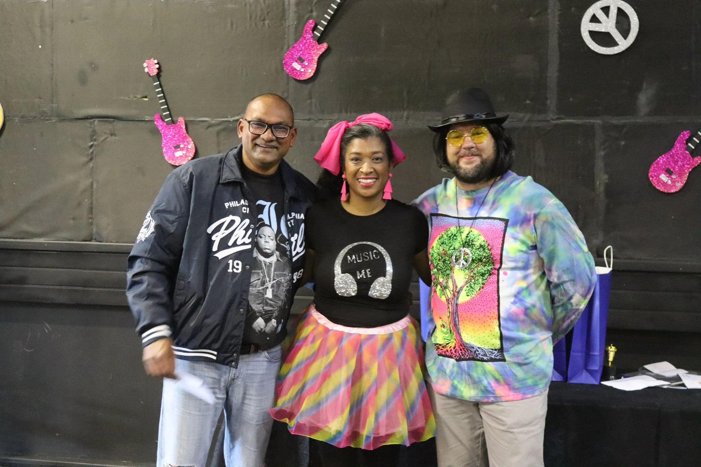 Three people standing together in front of a black wall decorated with pink glitter guitars and peace symbols, smiling at the camera.
