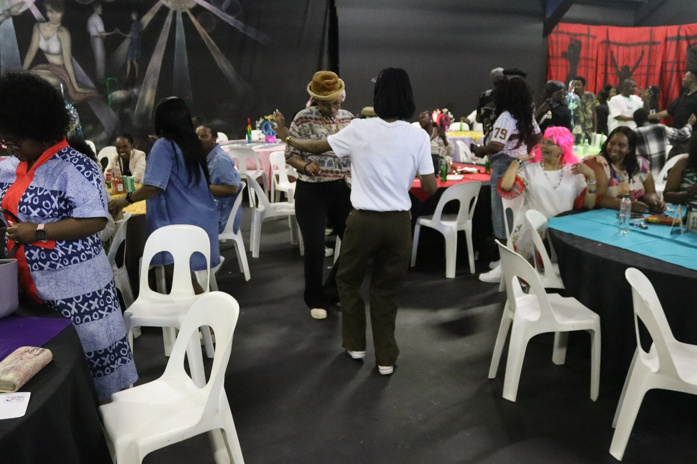 A diverse group of people gathered at round tables with colorful tablecloths in a festive indoor setting. The background features performances on a stage, including a woman with bright pink hair smiling and looking lively. Several people are dancing 