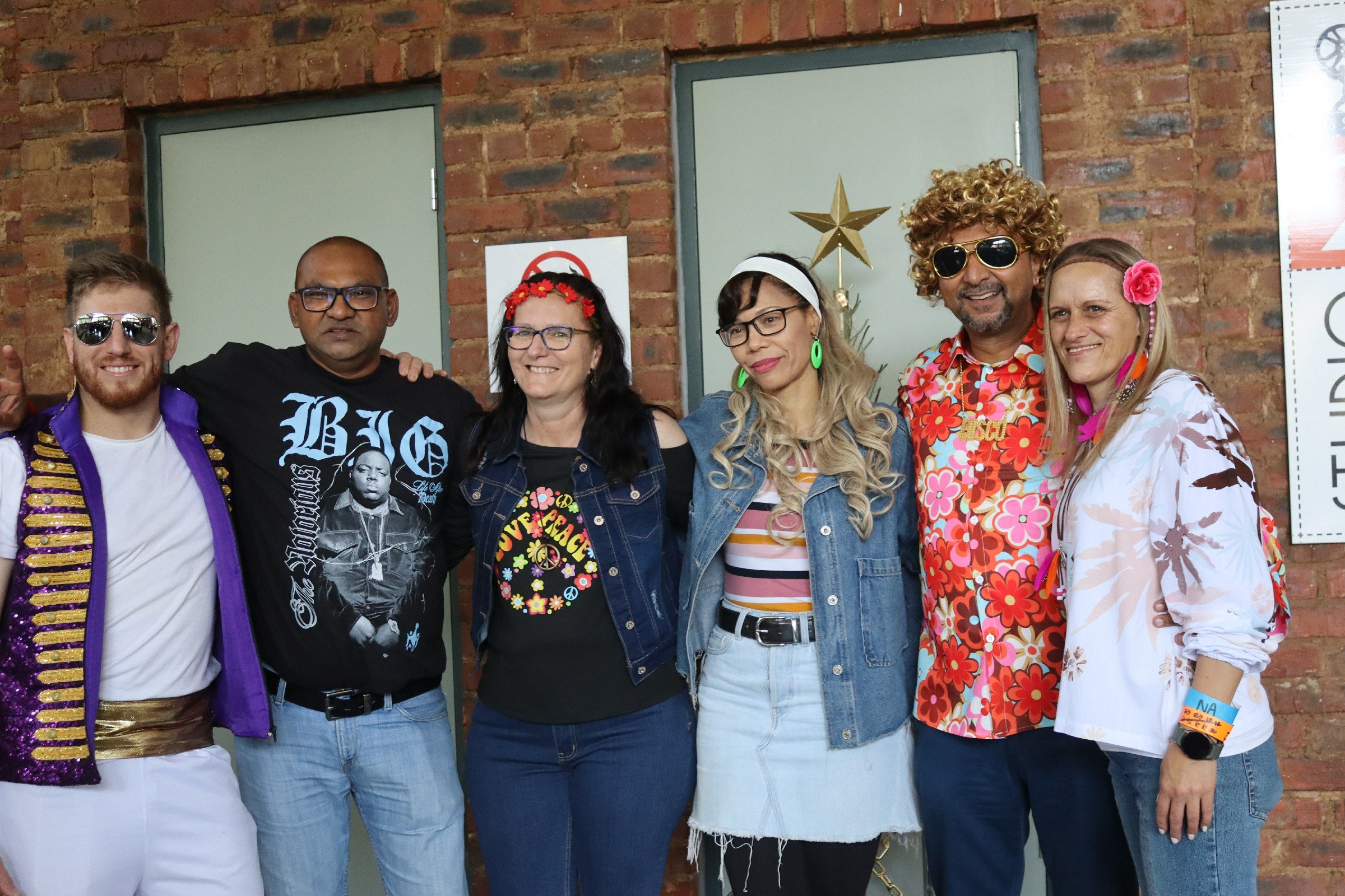 A group of seven people dressed in colorful, retro, 1960s-themed outfits posing in front of a brick wall with a Christmas tree and star decoration.