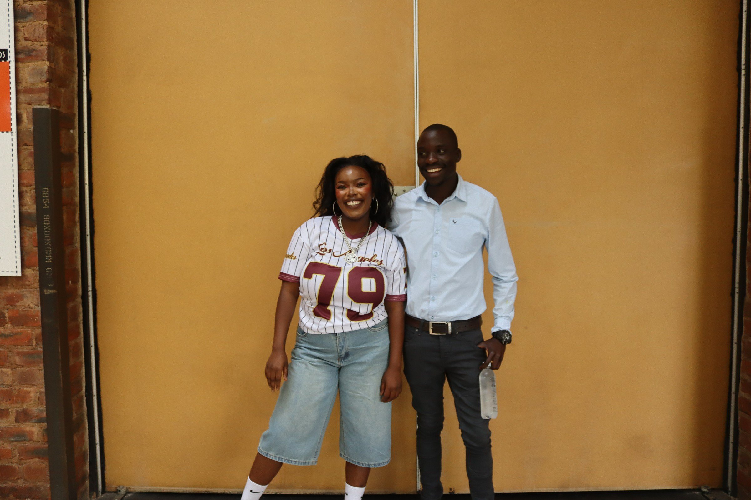 A woman and a man standing in front of a yellow backdrop, smiling for the camera. The woman is wearing a white jersey with maroon and yellow details and light blue jeans. The man is dressed in a white button-up shirt and dark pants, holding a phone i