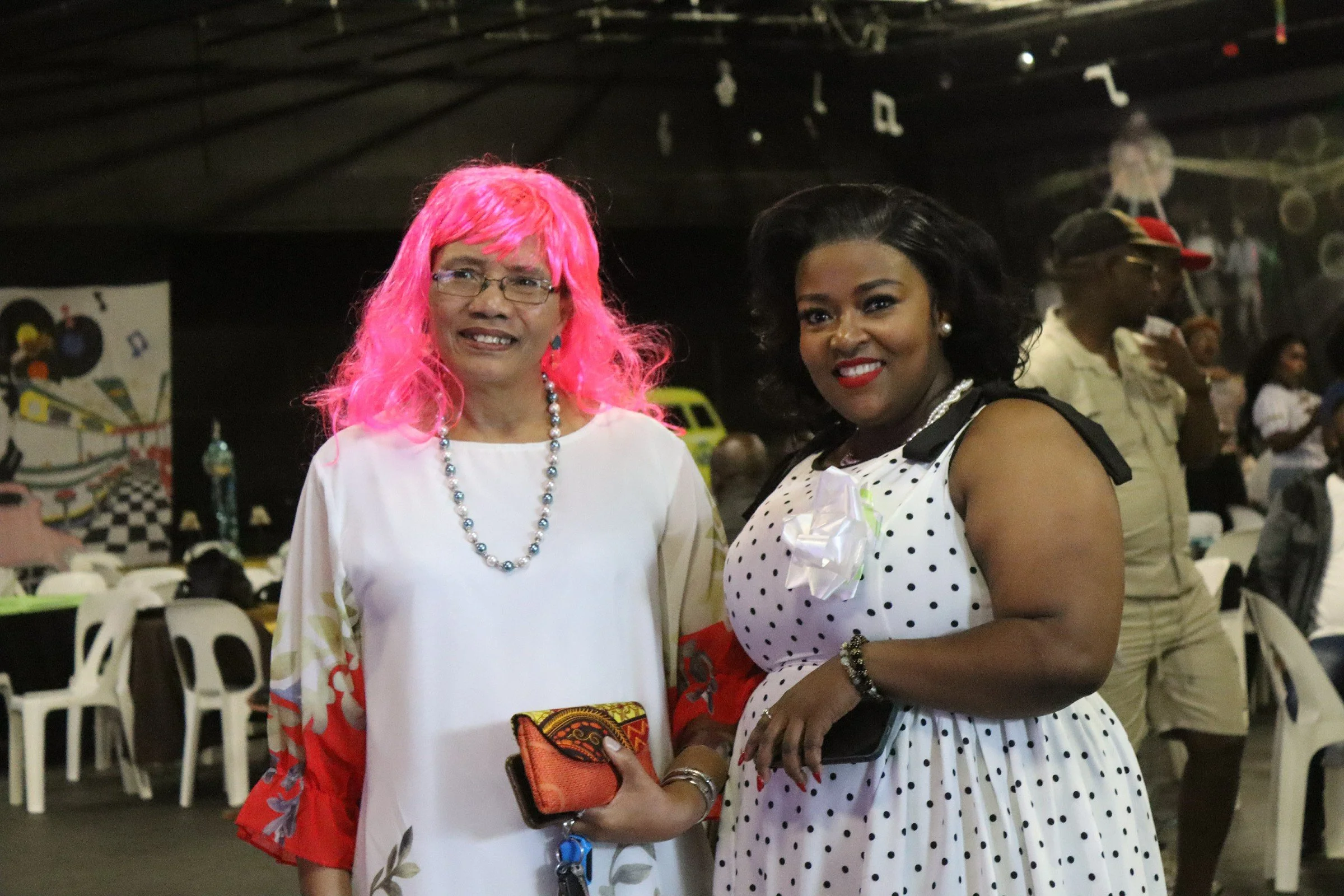 Two women standing together in an indoor event space, both smiling at the camera. The woman on the left has pink hair, glasses, and is wearing a white dress with a colorful pattern and a beaded necklace. The woman on the right has dark, curly hair, r
