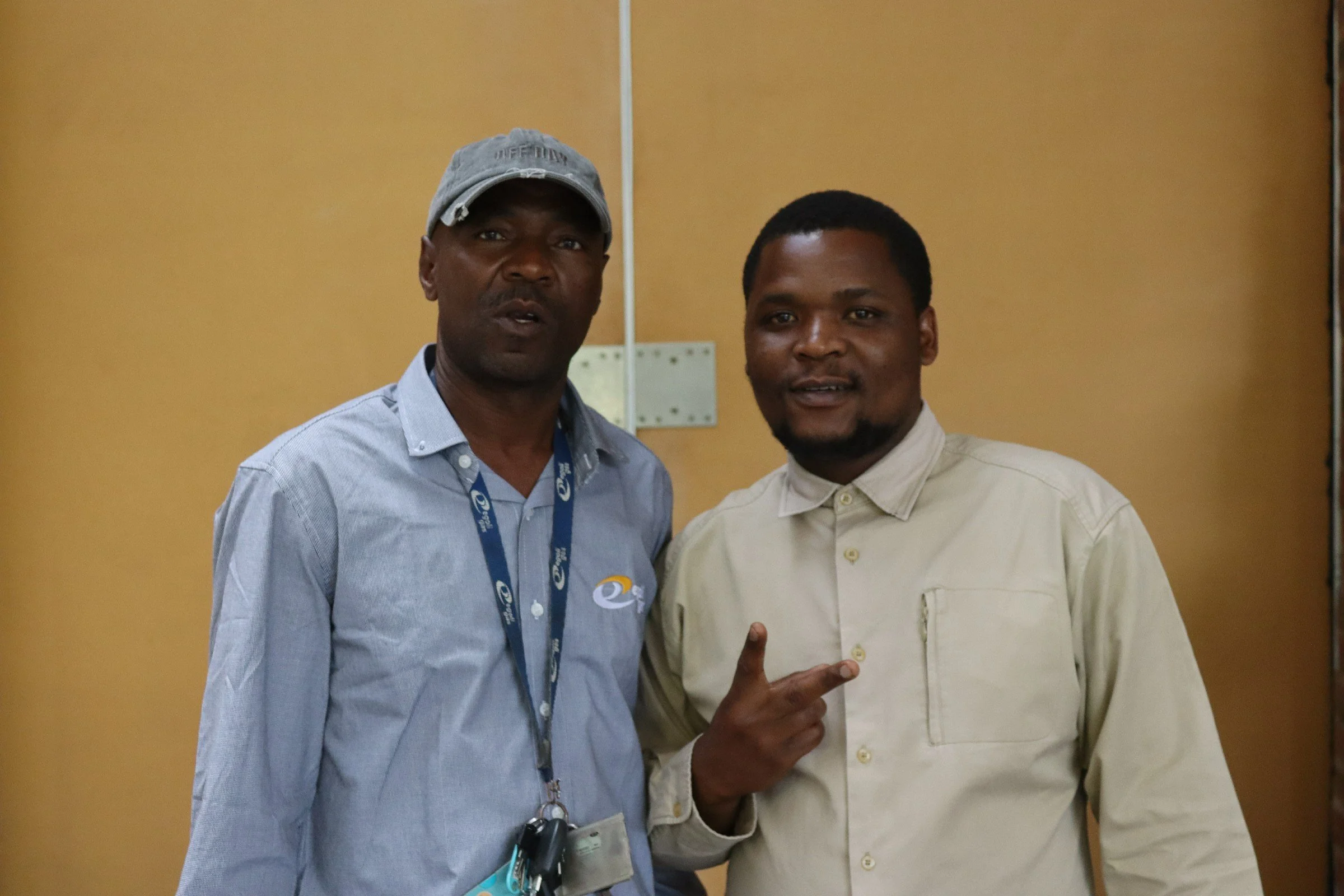 Two men standing in front of a plain beige door, one wearing a baseball cap and the other showing a peace sign.