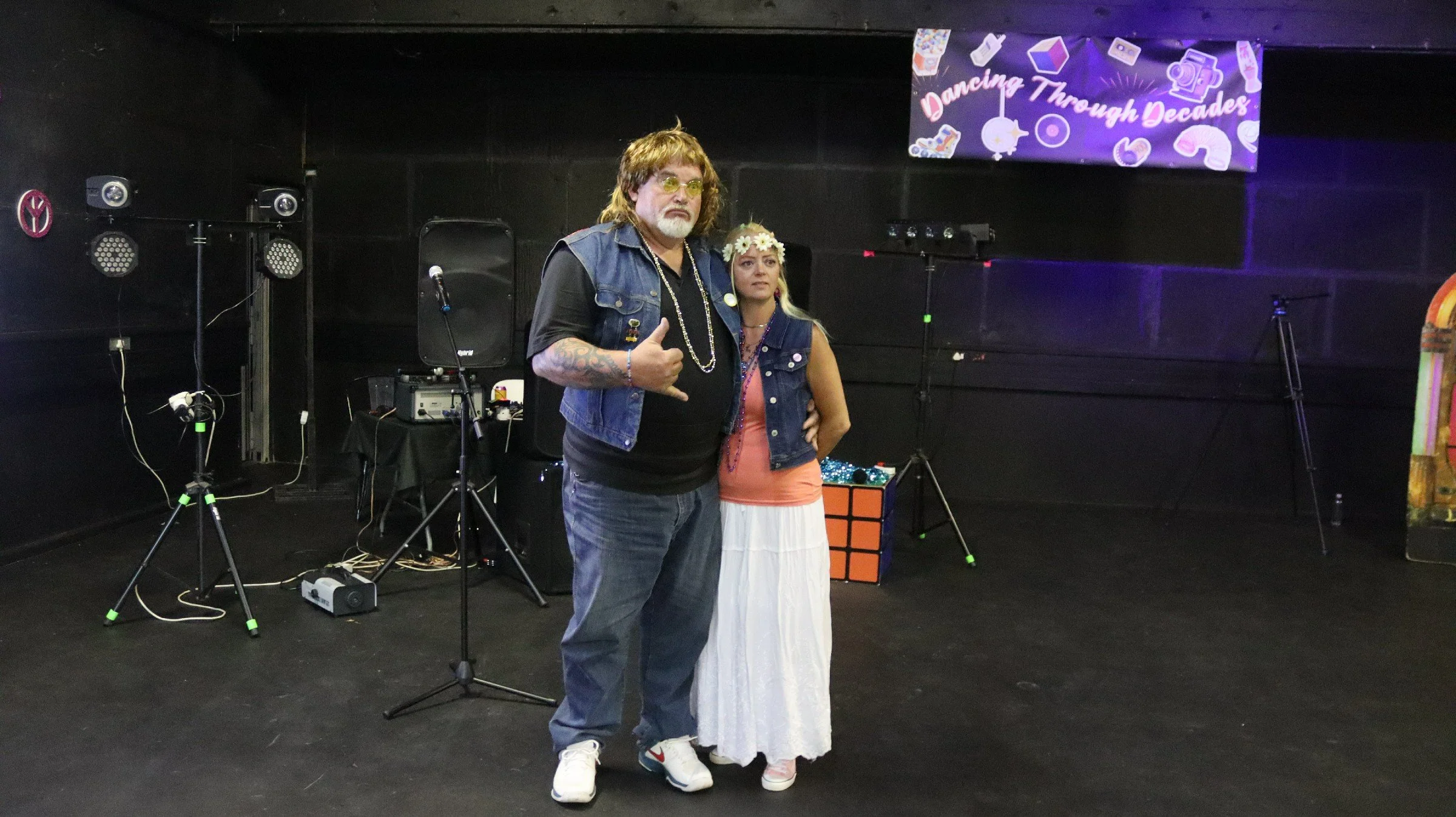 Two people, a man and a woman, standing on a stage with audio equipment and a colorful banner. The man has long hair, glasses, tattoos, and is making a gesture with his hand. The woman, wearing a flower crown and a white skirt, is standing next to hi
