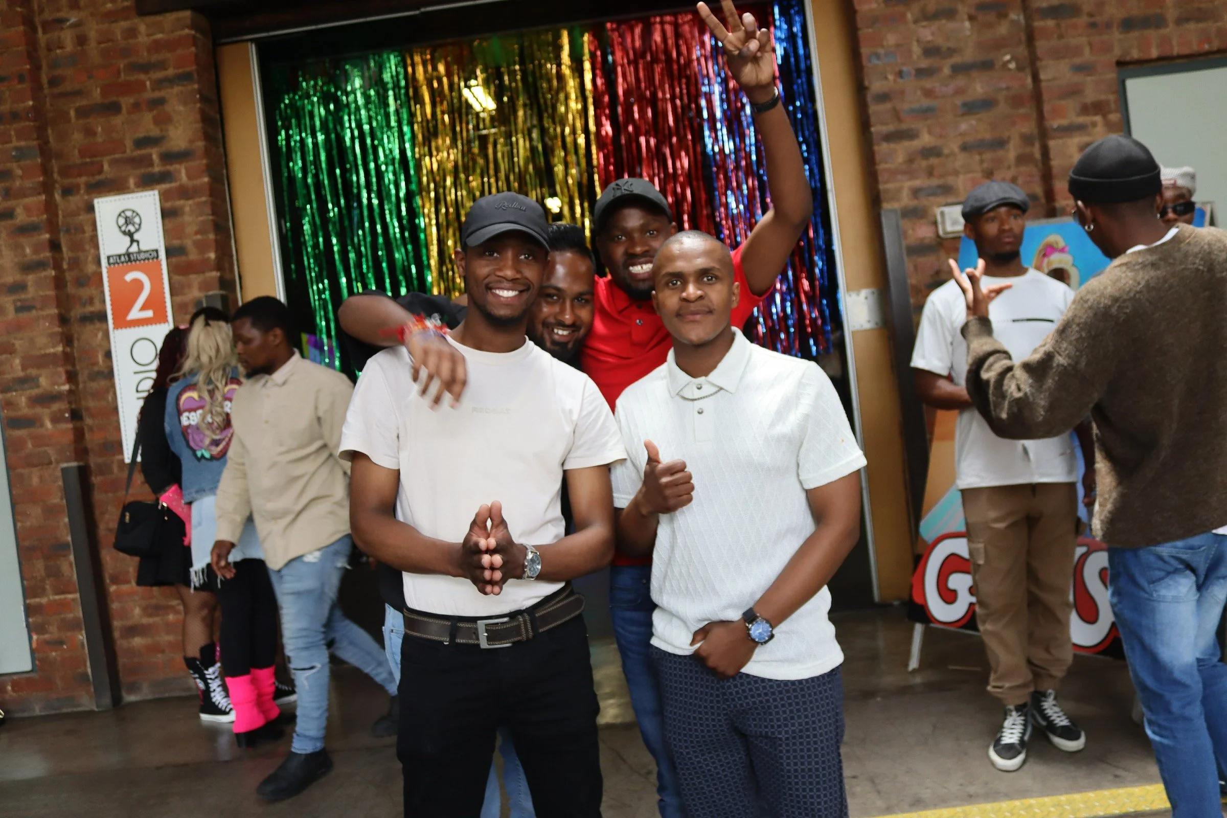 A group of young men smiling and posing together in front of a colorful, metallic curtain at an indoor event, with some making peace signs and others with their arms around each other.