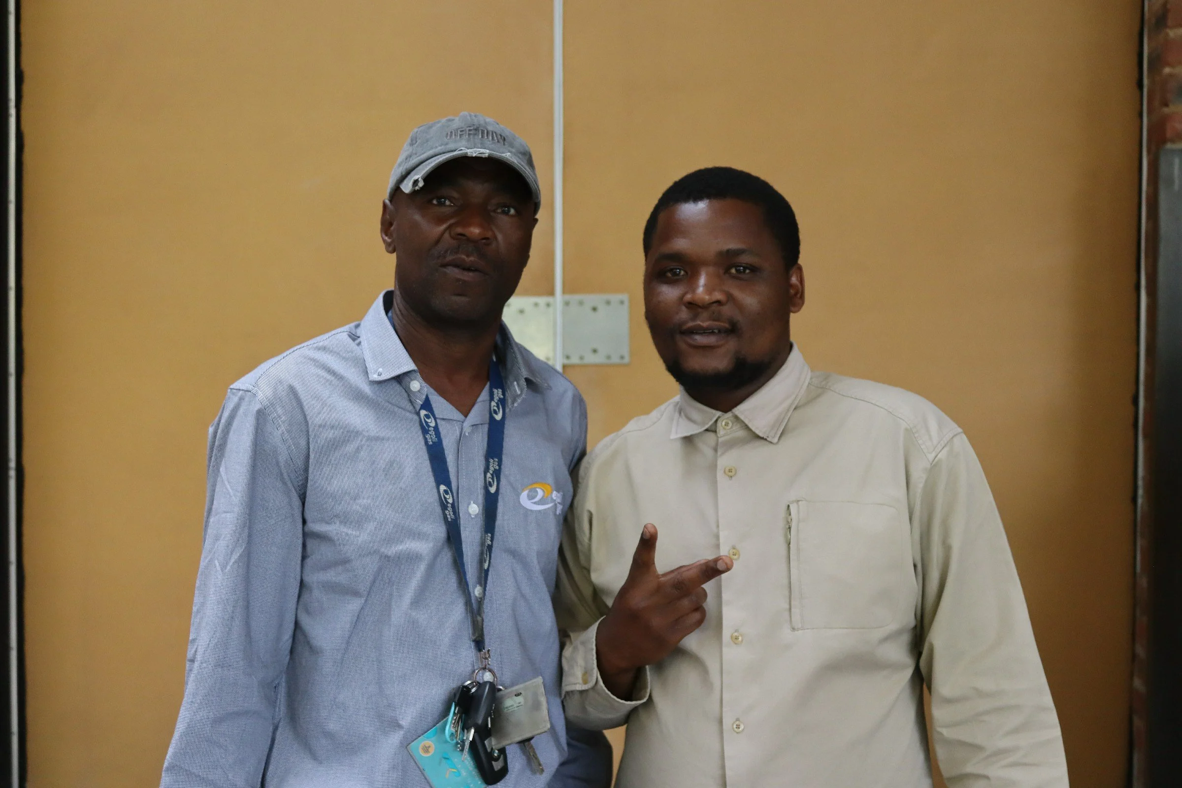 Two men standing side by side, one wearing a light blue dress shirt, a gray cap, and lanyard with ID badge, and the other wearing a cream-colored shirt. The man on the right is making a peace sign gesture with his fingers. They are inside a room with