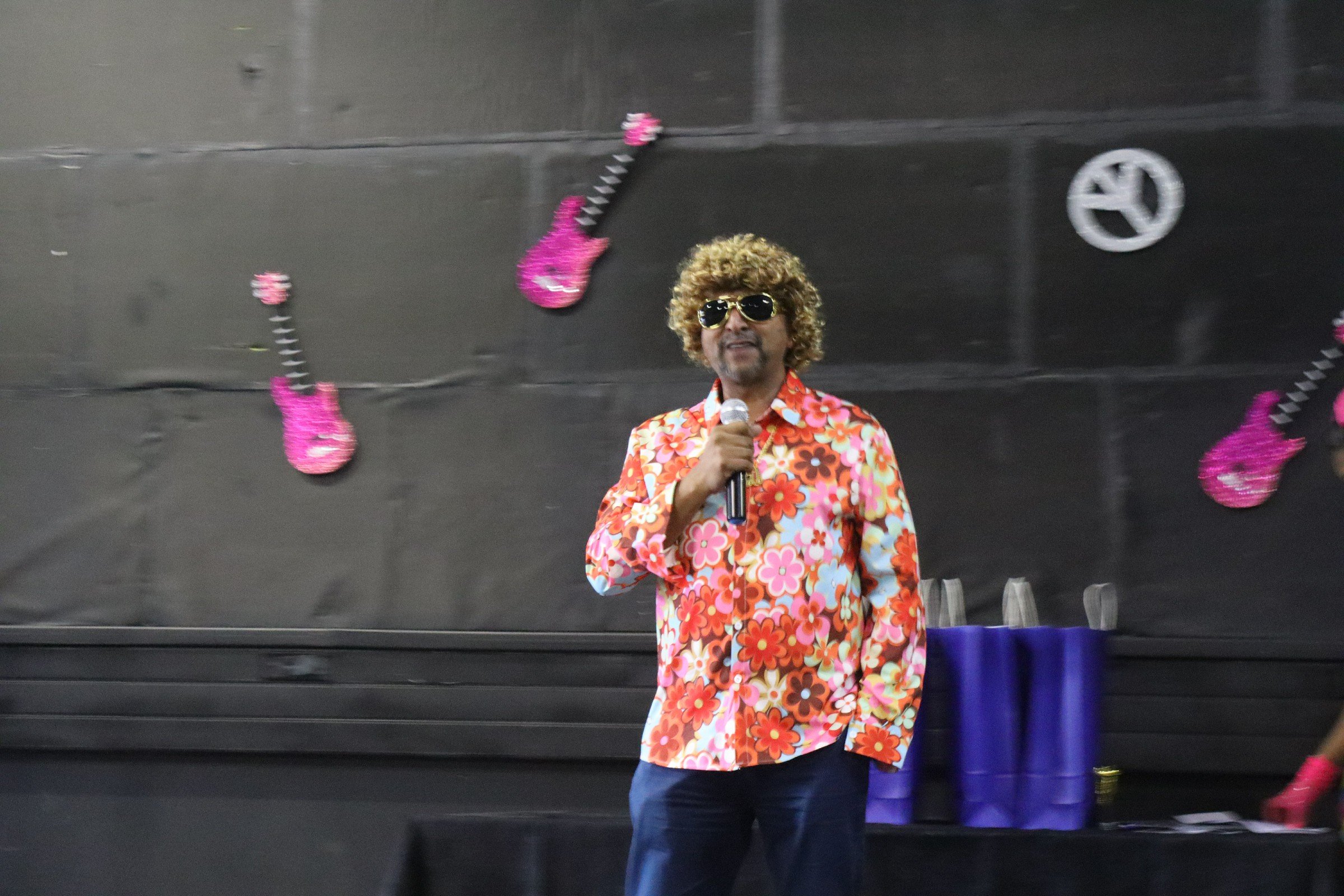 A man with curly hair and sunglasses wearing a colorful floral shirt, holding a microphone, standing in front of a black wall decorated with pink guitar-shaped ornaments and a no-smoking sign.