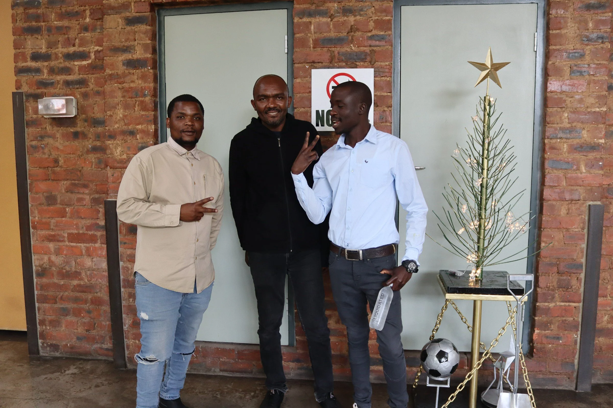 Three men standing in front of a brick wall, posing for a photo. One man is making a peace sign, and they are dressed in casual and semi-formal clothing. To the right is a small decorated Christmas tree with a gold star on top, next to a soccer ball.