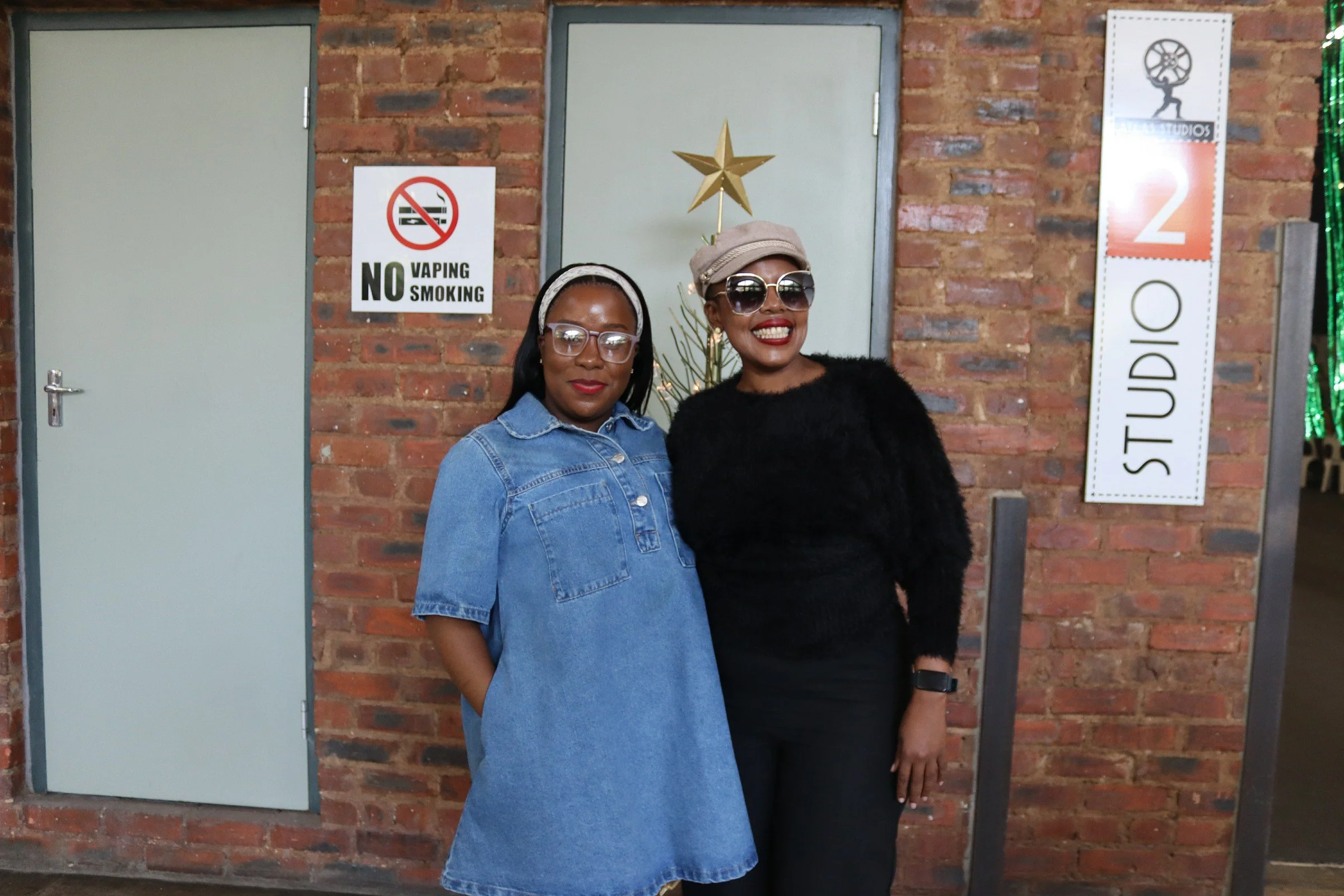 Two women standing in front of a brick wall with a 'No Smoking' sign and a tall, decorated Christmas tree. One woman is wearing a denim dress and glasses, the other is wearing a black fuzzy top, a beige hat, and sunglasses. The woman in black is smil