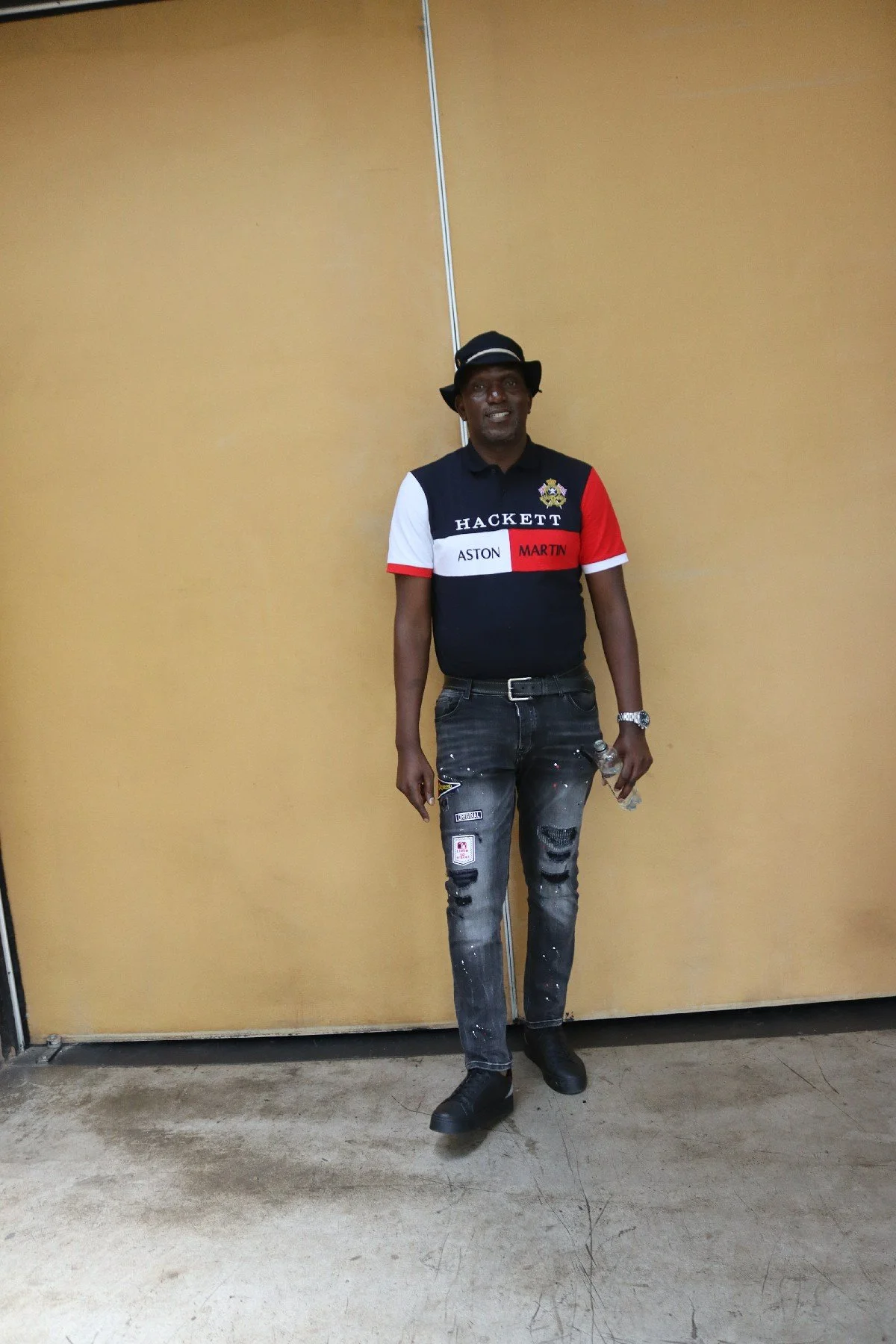 Man standing in front of a beige door, wearing a black hat, a colorful shirt with patches, distressed jeans, and holding a water bottle.