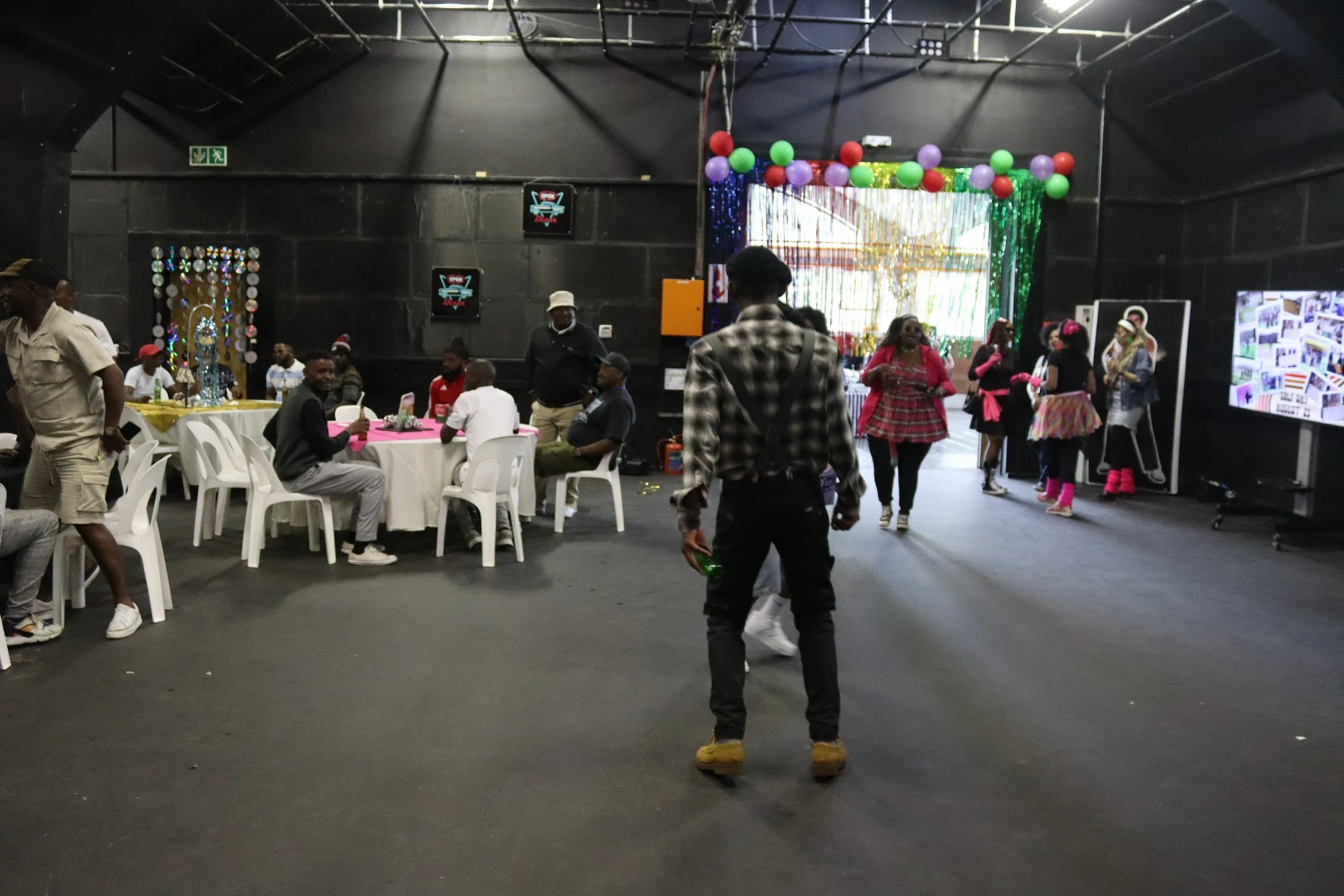 Interior of a festive event space with people gathering, decorated with colorful balloons and streamers, some people sitting at tables, and a person walking in the foreground.