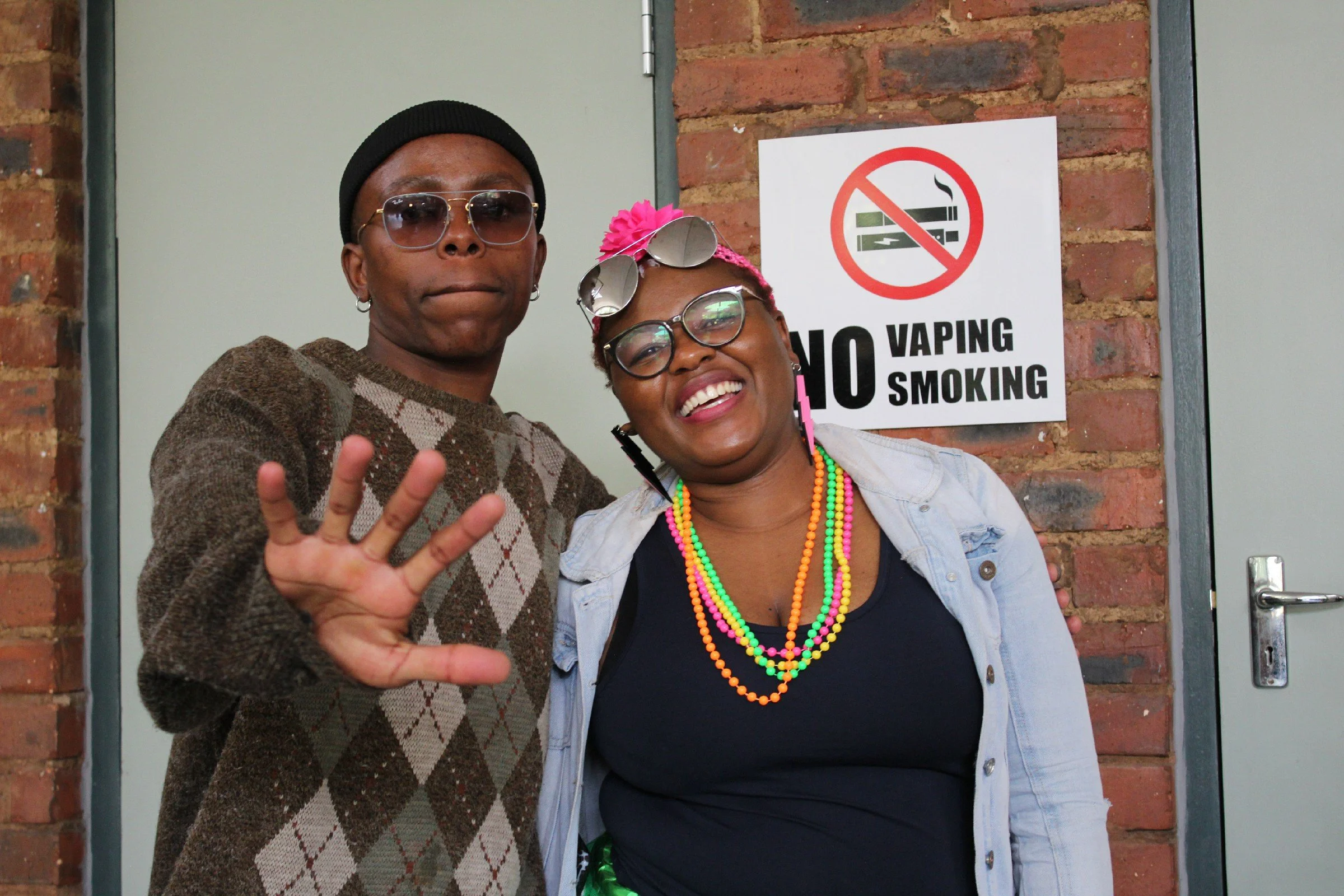 Two people standing together in front of a brick wall with a no vaping or smoking sign; the person on the left is wearing a brown argyle sweater, black beanie, and sunglasses, making a peace sign; the person on the right is smiling, wearing glasses, 