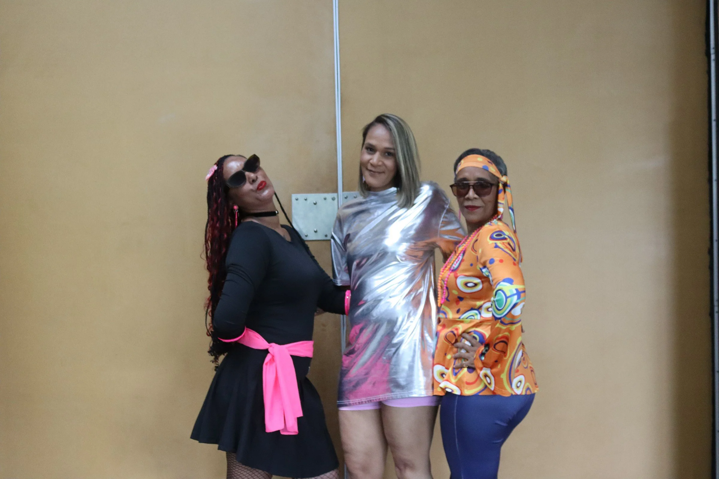 Three women standing together in front of a beige wall, dressed in colorful and stylish clothing, posing confidently with smiles.