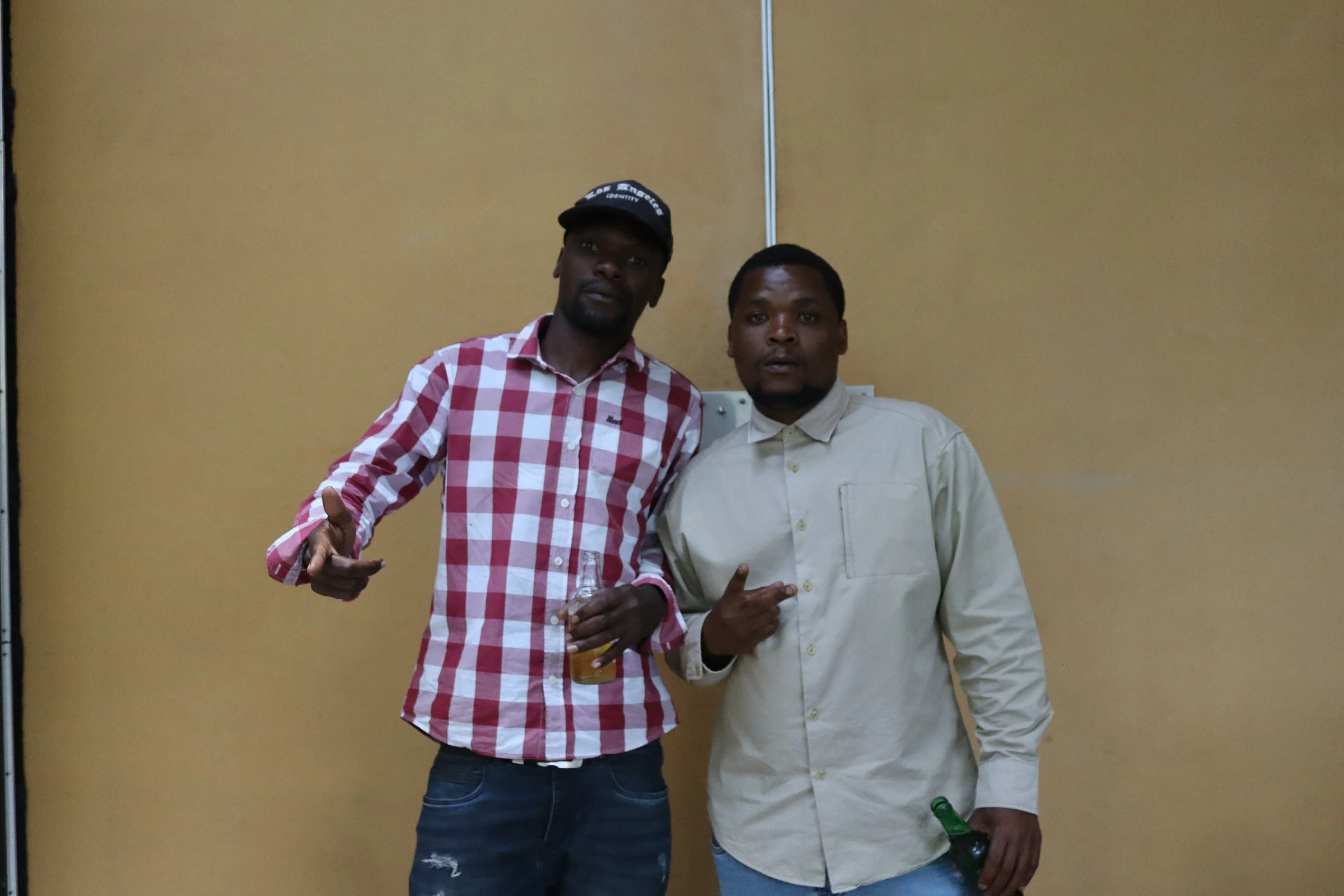 Two men standing together in front of a yellow wall, one holding a drink and the other holding a beer bottle, both making hand gestures.