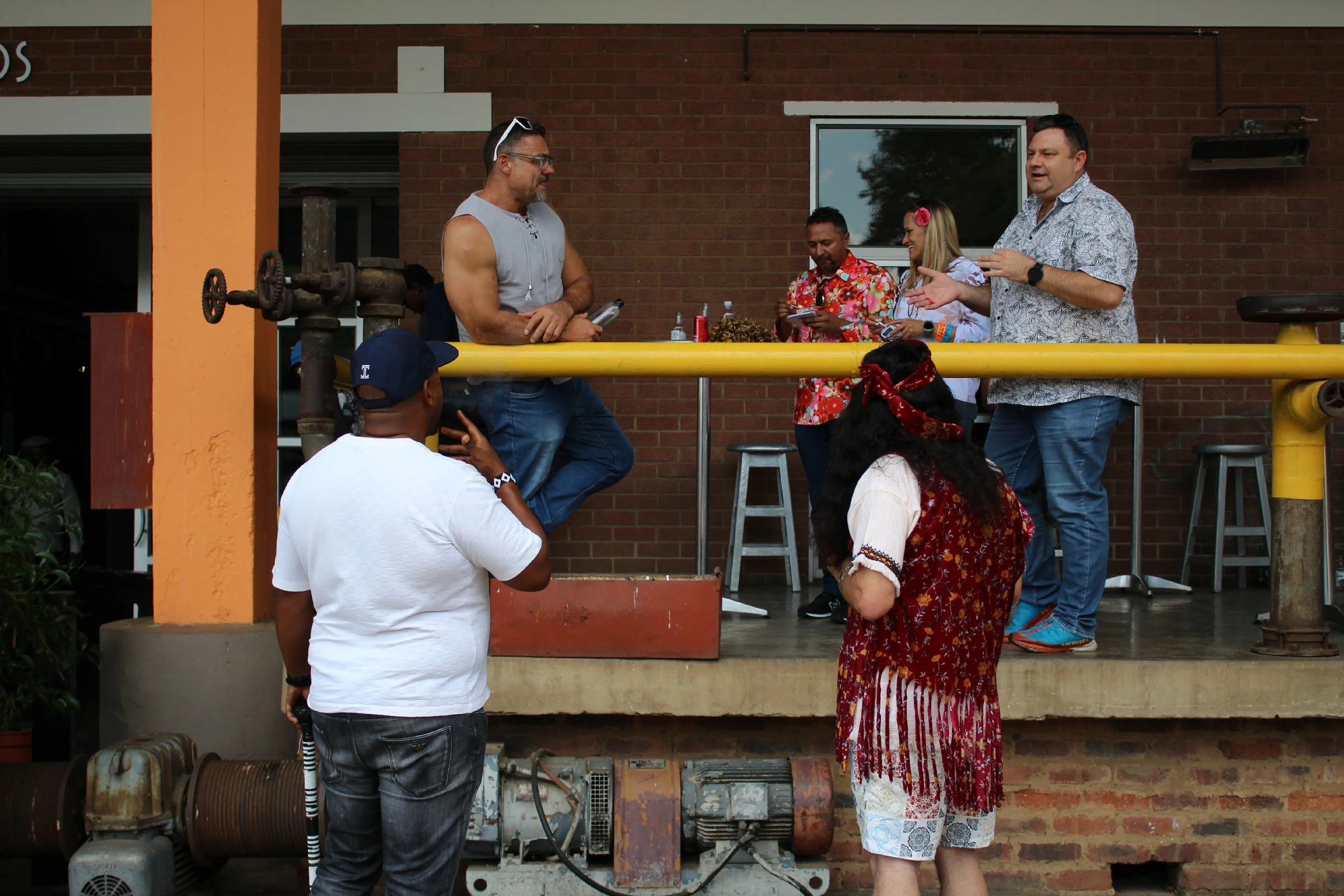 A group of people attending a tropical-themed outdoor event. Two men are standing near a yellow pipe, one with a sleeveless shirt and the other with a printed short-sleeve shirt. Two women are also present, one in a floral dress and the other with a 