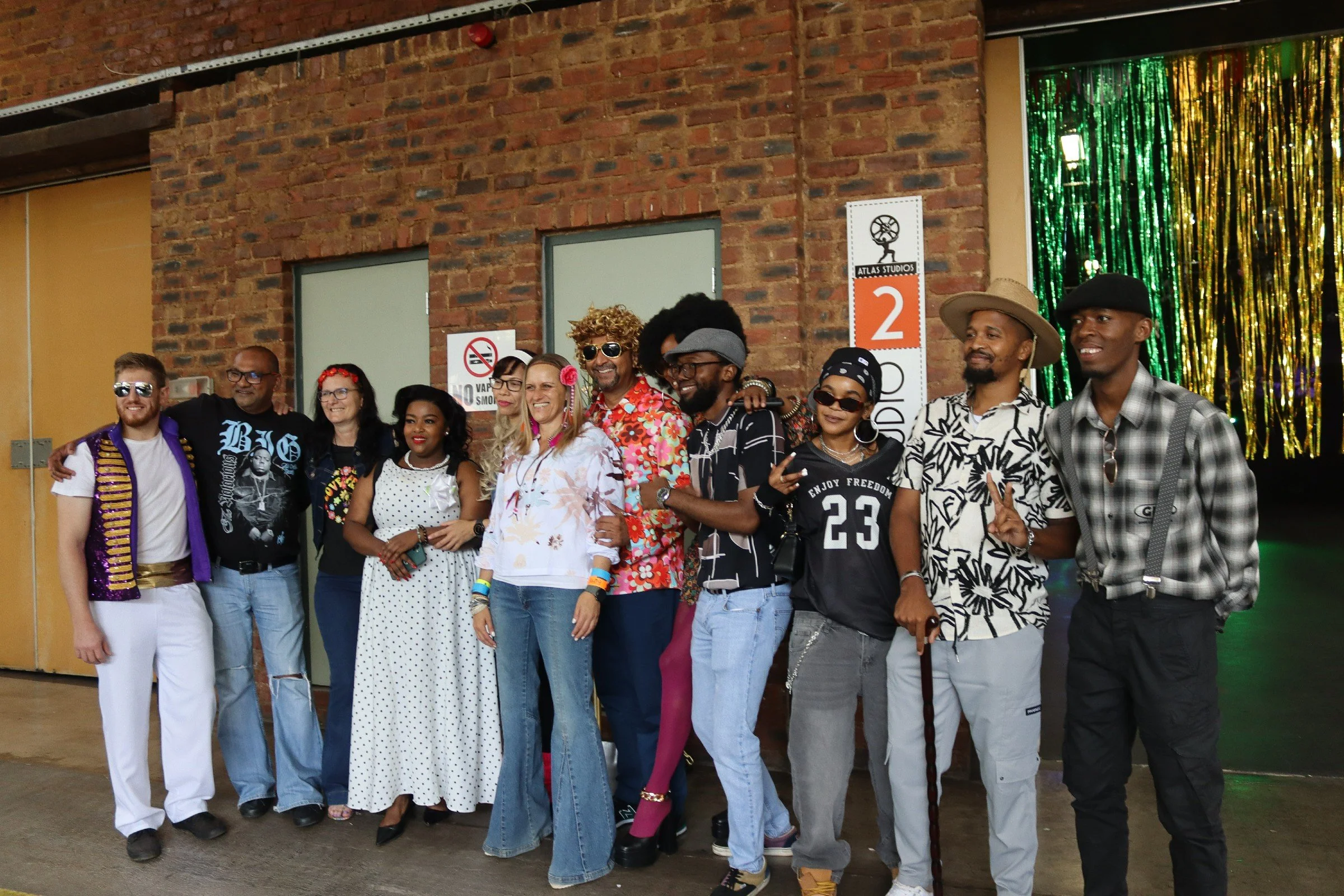 A diverse group of people smiling and posing together in front of a brick wall, with a colorful backdrop and signage that reads 'Atlas Studios' and '2'.