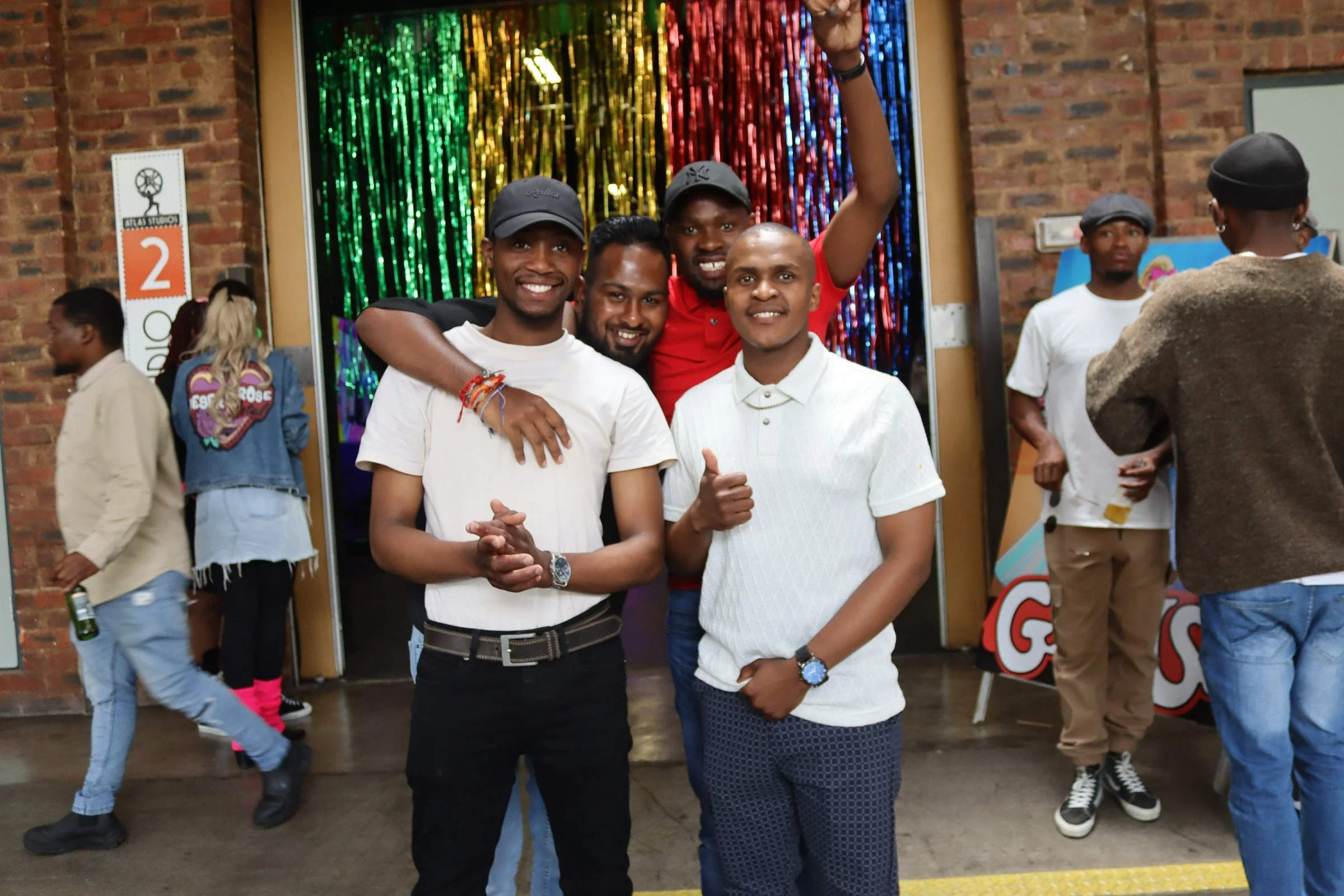 Group of five young men posing for a photo at an indoor event with a colorful, shiny backdrop and brick walls. Four are smiling at the camera, one has his arm around another, and one man is giving a thumbs-up.