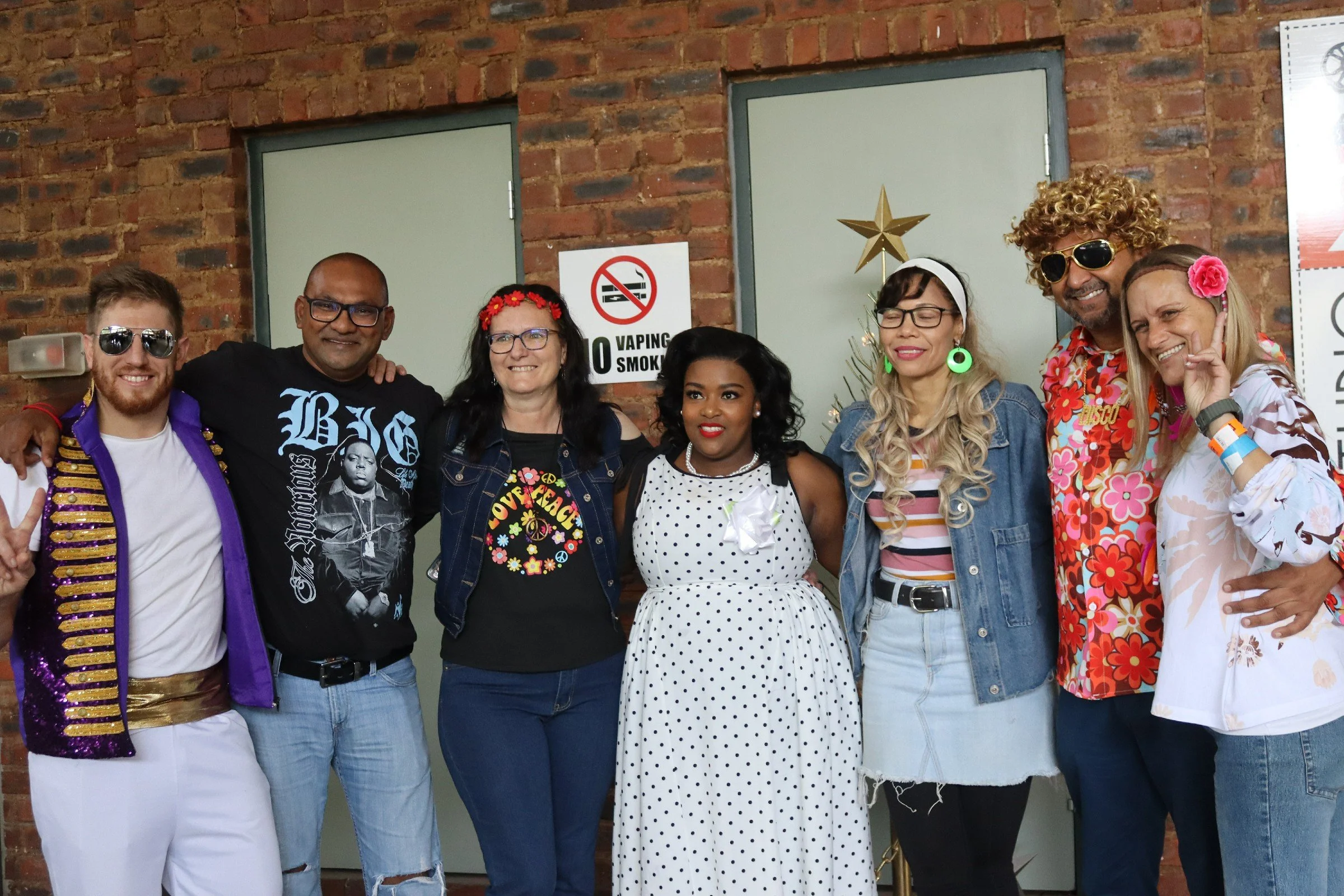 A diverse group of eight people smiling and posing together indoors, standing in front of a brick wall with a 'No Vaping' sign, some wearing colorful and retro clothing, accessories, and glasses.
