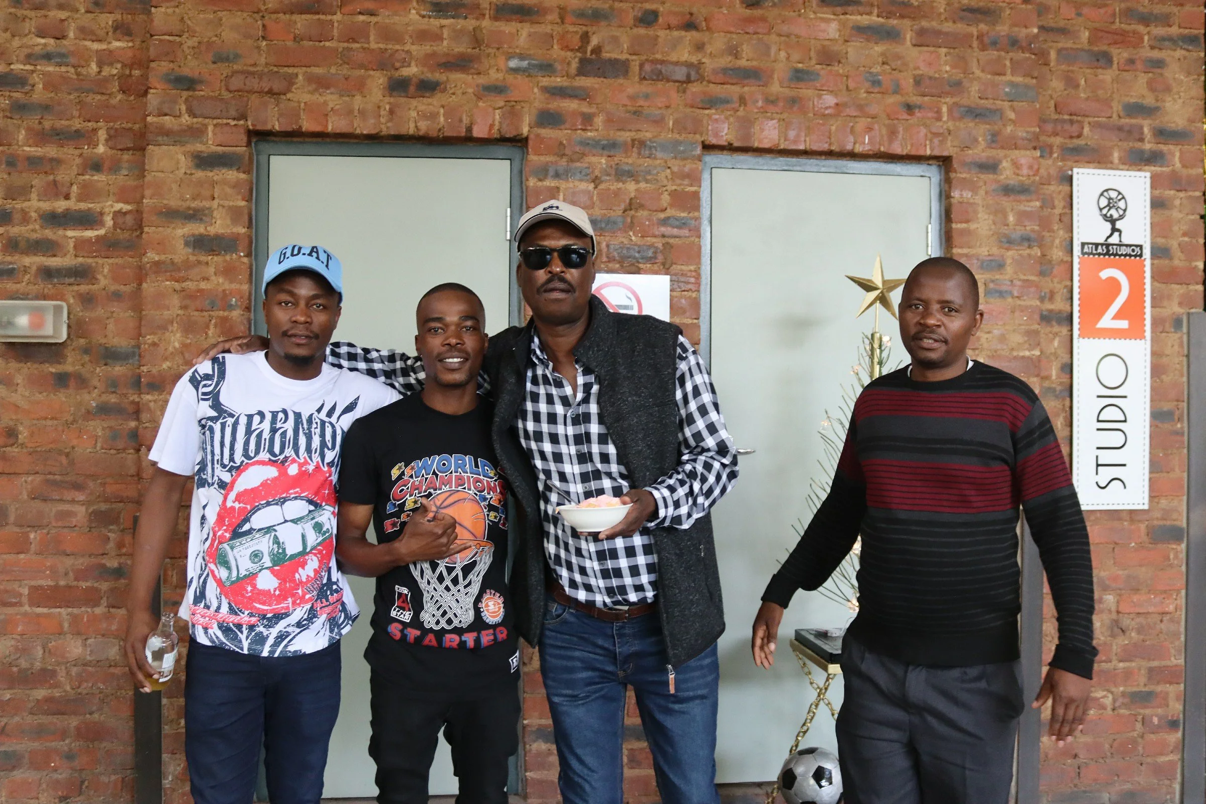 Four men standing in front of a brick wall, posing for a photo. One man is holding a bowl, and another man has his arm around his shoulder. There is a small Christmas tree and a soccer ball at the bottom right corner. A sign with the number 2 and the